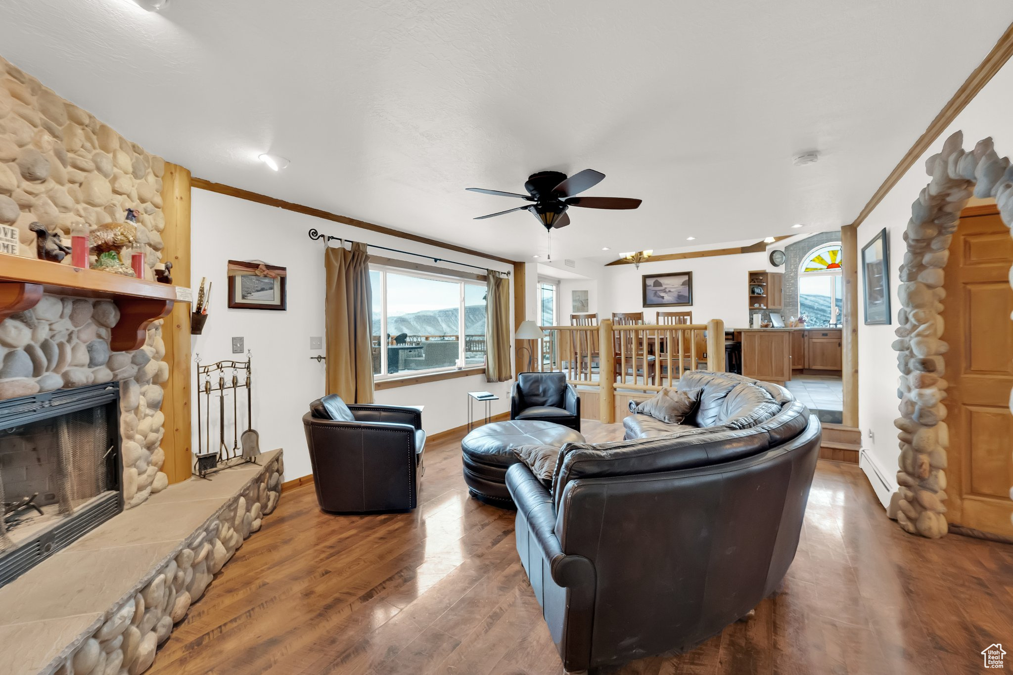 Living room featuring ceiling fan, a baseboard heating unit, wood-type flooring, ornamental molding, and a stone fireplace