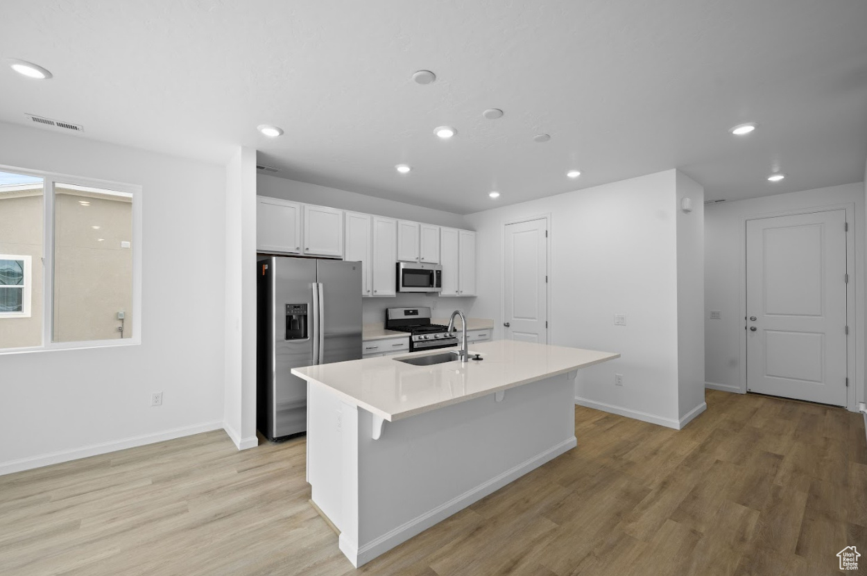 Kitchen with sink, light LVT style flooring, appliances with stainless steel finishes, a kitchen island with sink, and white cabinetry with hardware