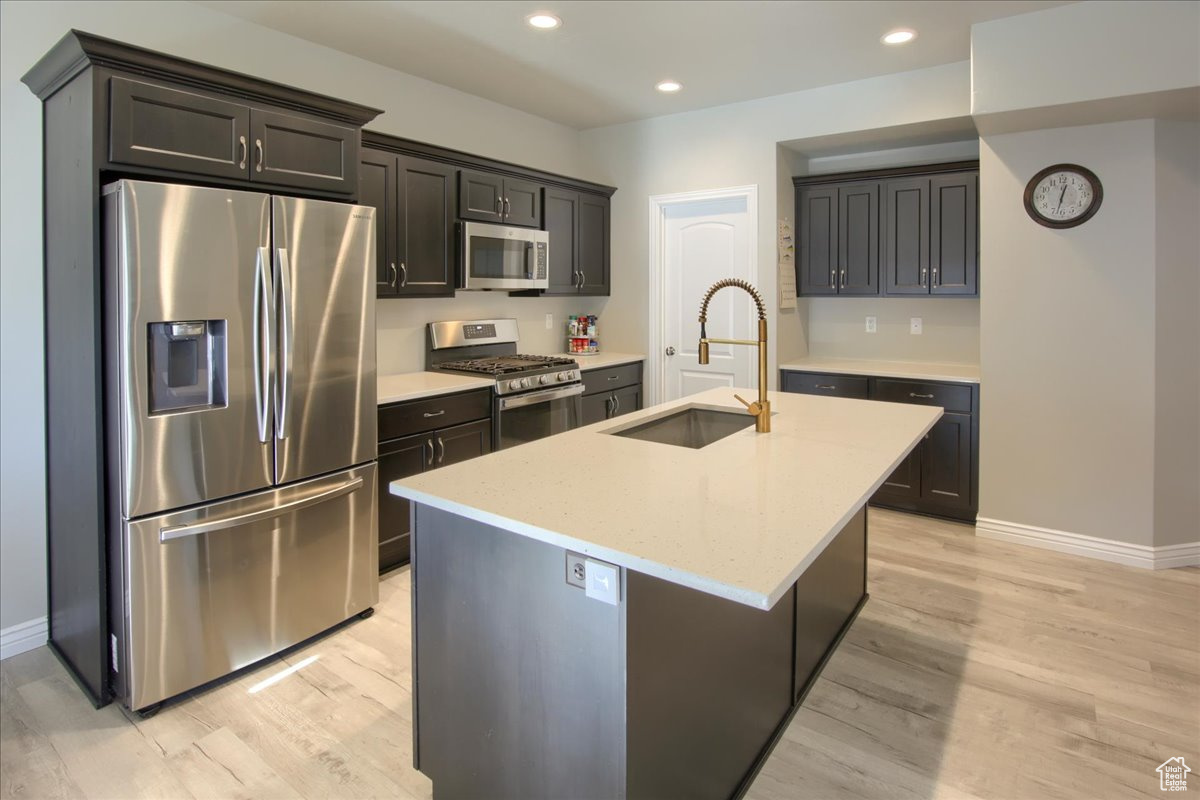 Kitchen with sink, light stone counters, a center island with sink, light hardwood / wood-style flooring, and appliances with stainless steel finishes