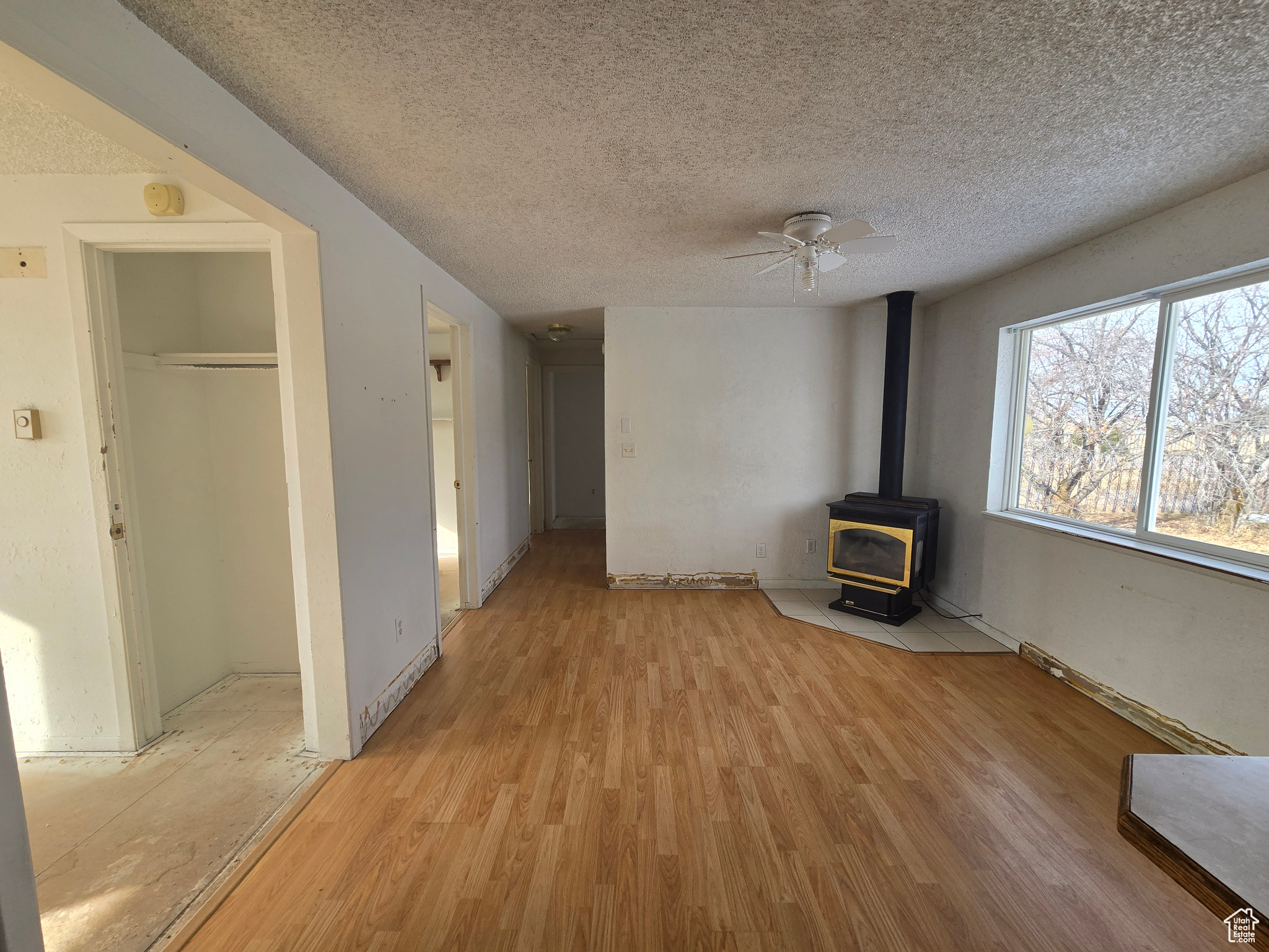 Unfurnished living room featuring ceiling fan, a wood stove, a textured ceiling, and light hardwood / wood-style floors