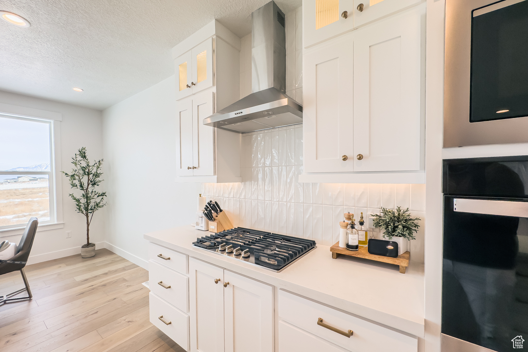 Kitchen featuring black oven, stainless steel gas stovetop, white cabinets, wall chimney range hood, and light wood-type flooring