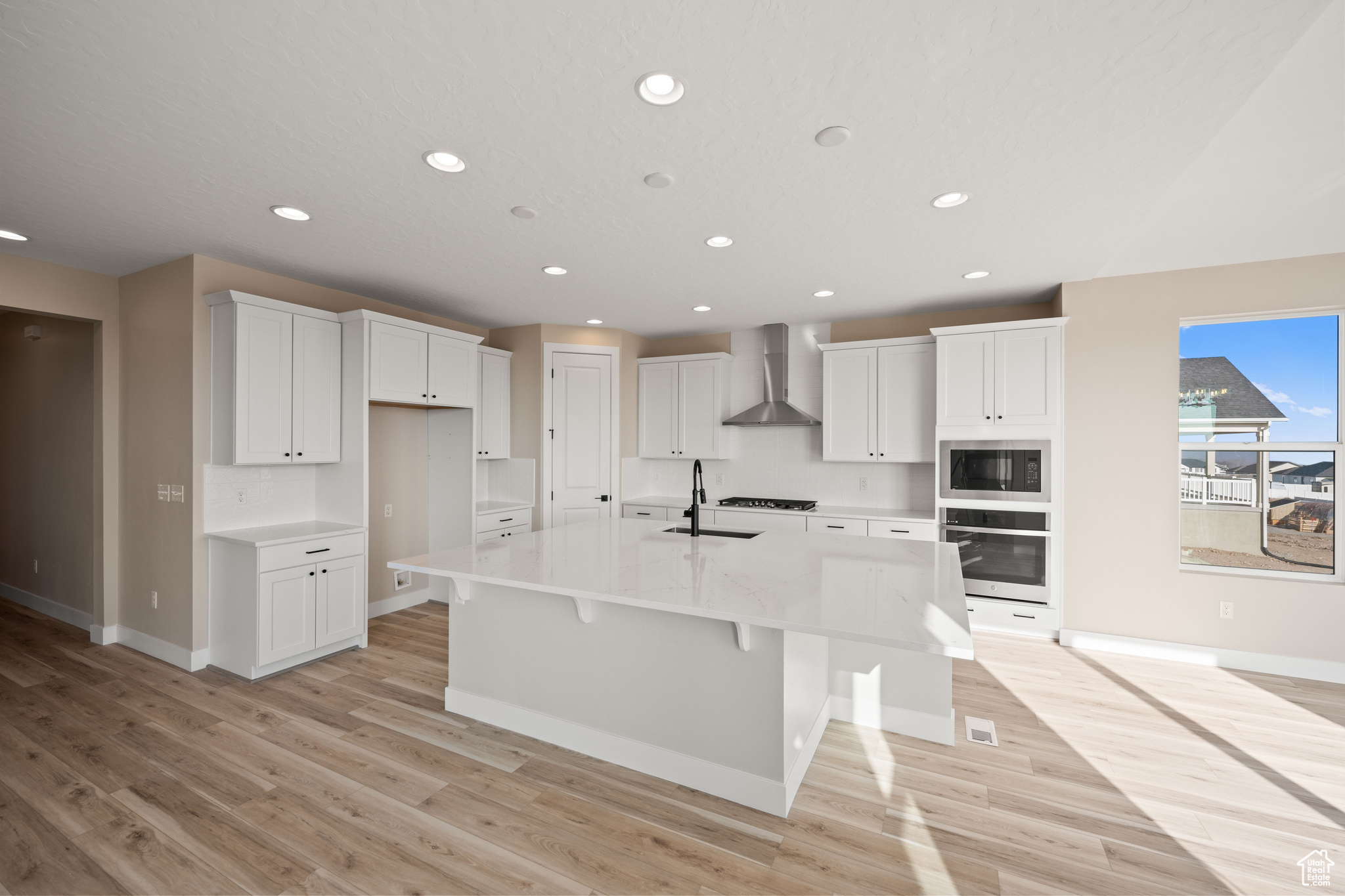 Kitchen featuring built in microwave, white cabinetry, an island with sink, oven, and wall chimney range hood