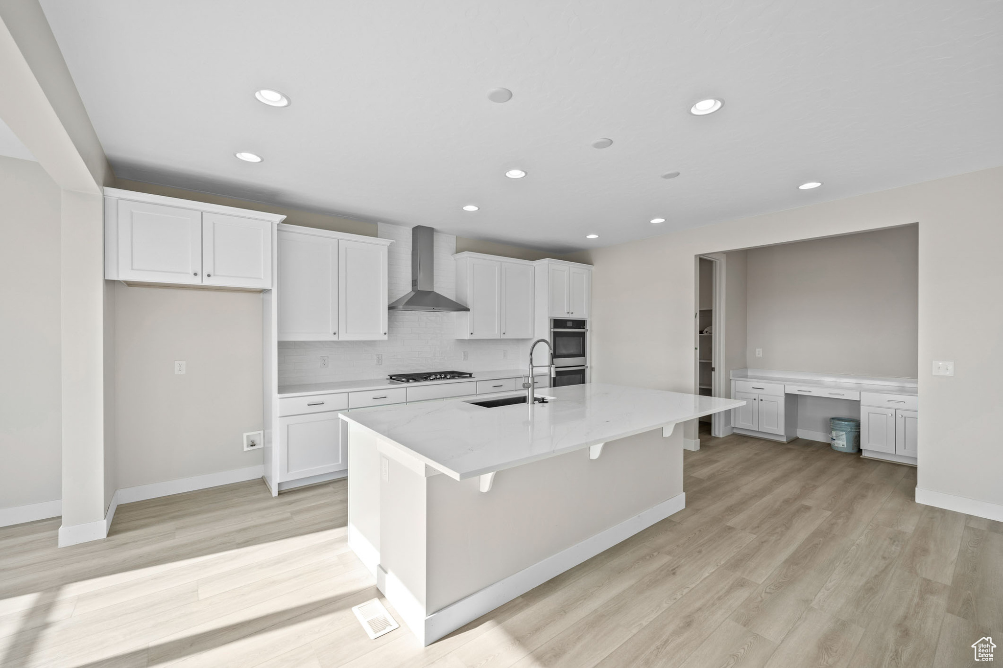 Kitchen with built in desk, white cabinetry, sink, a kitchen island with sink, and wall chimney range hood