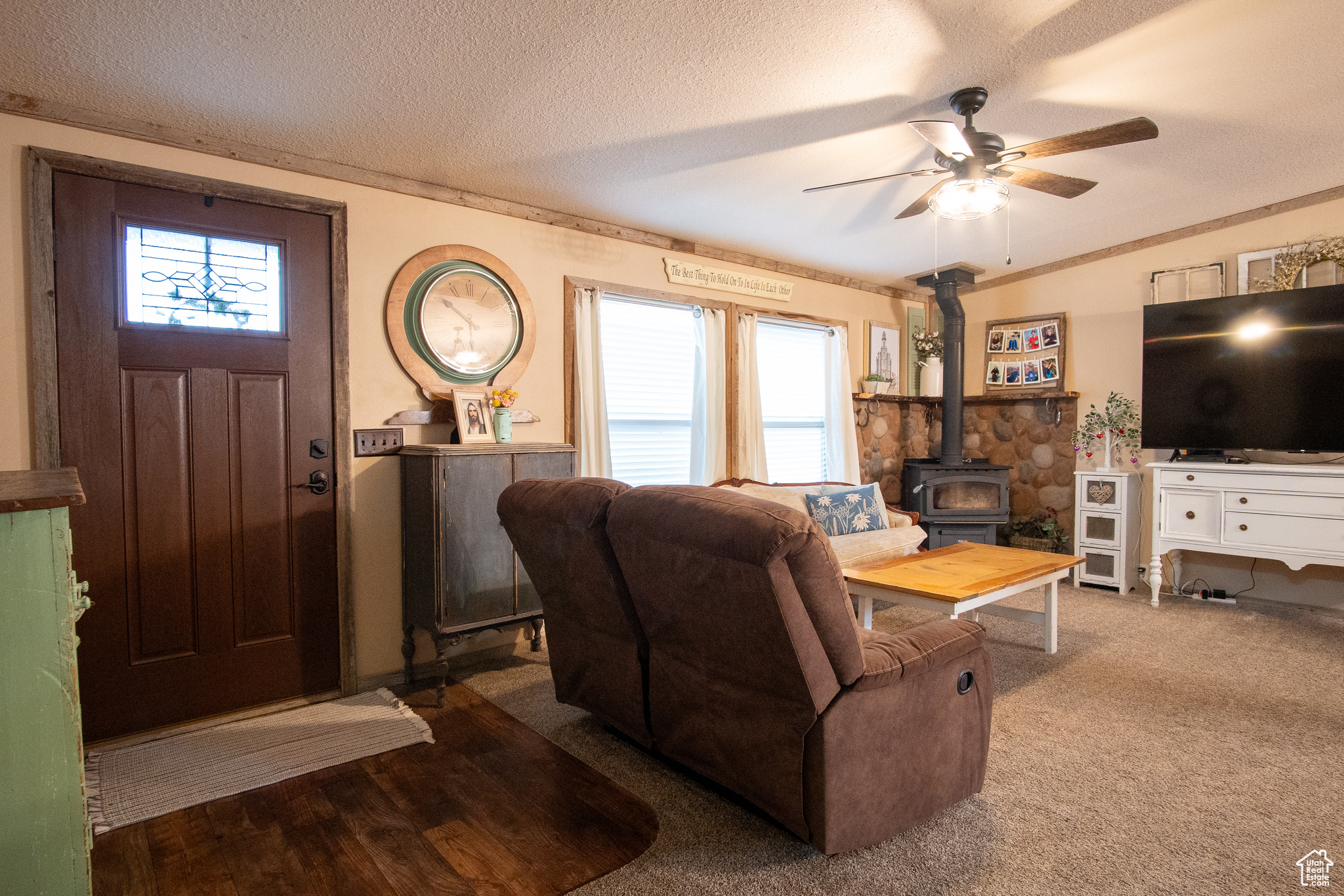 Carpeted living room with ceiling fan, plenty of natural light, a textured ceiling, and a wood stove