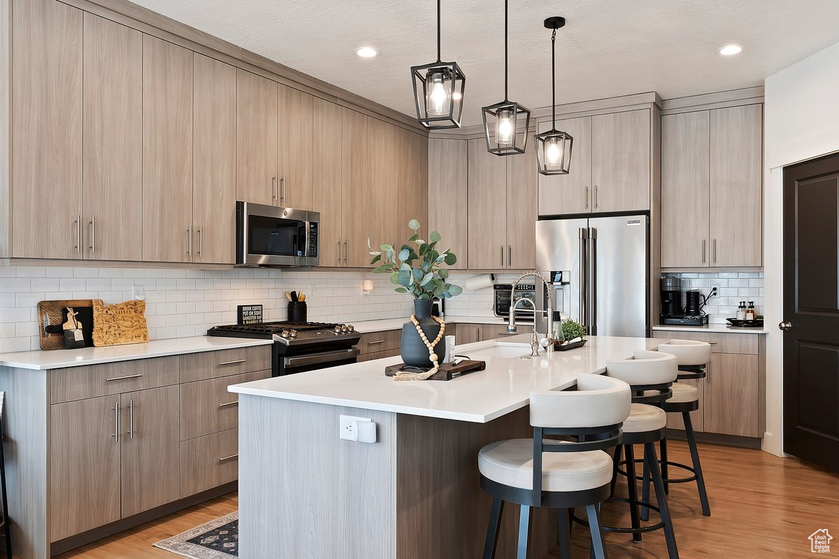Kitchen with light hardwood / wood-style flooring, hanging light fixtures, stainless steel appliances, a center island with sink, and light brown cabinets
