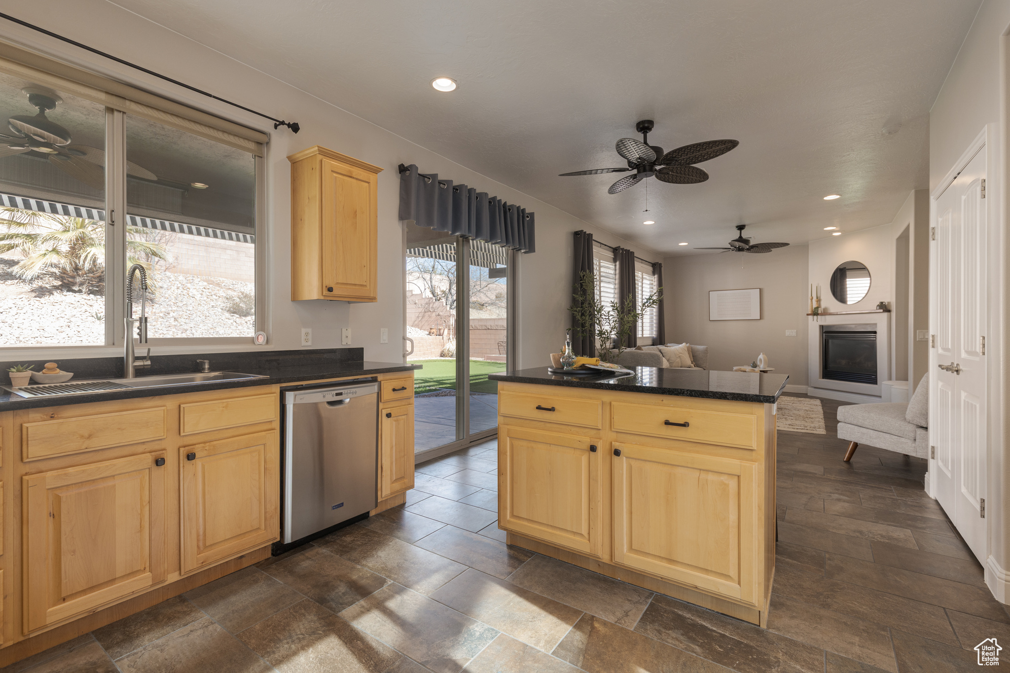 Kitchen featuring dark countertops, light brown cabinetry, open floor plan, stainless steel dishwasher, and a sink