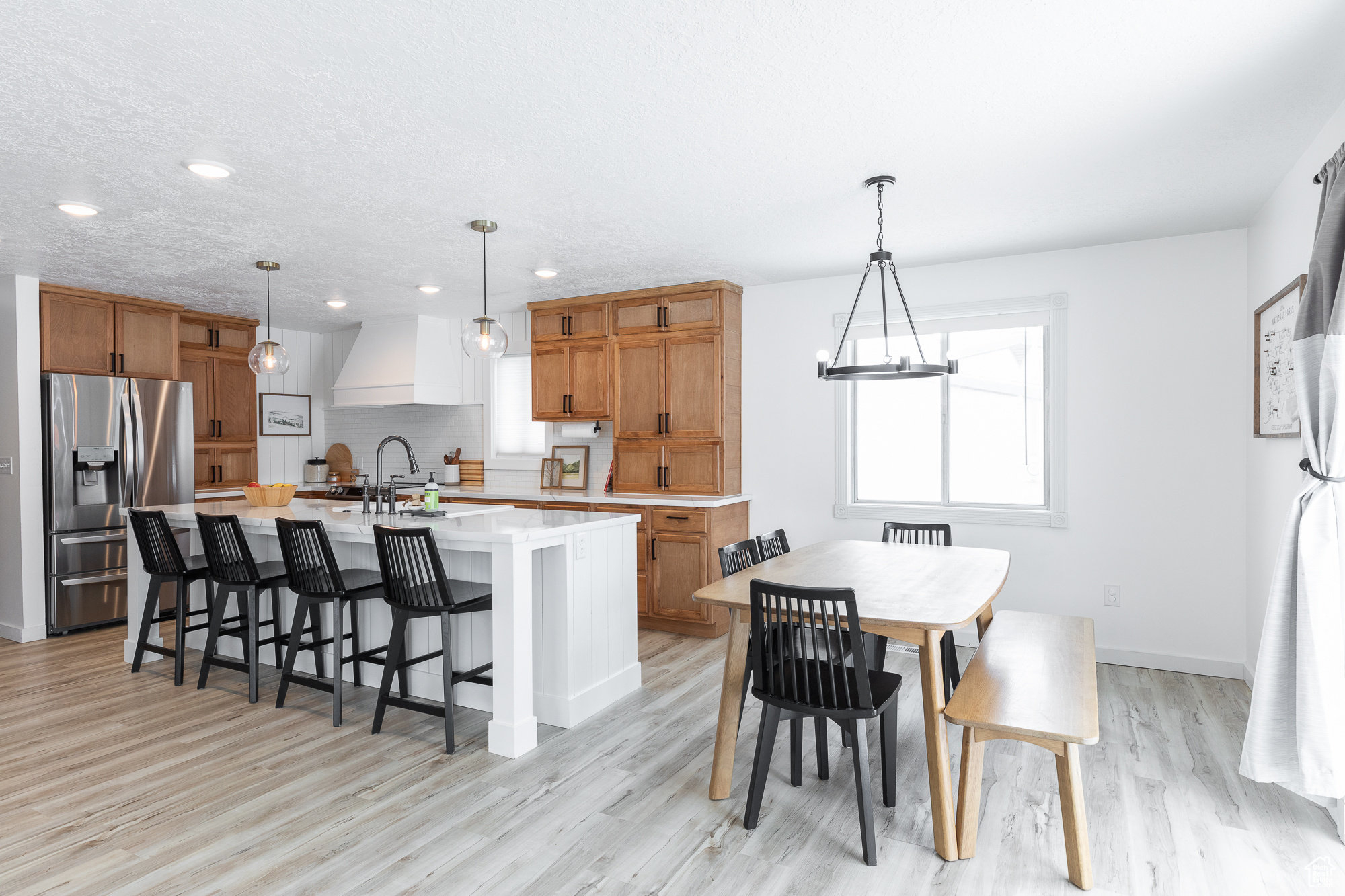 Kitchen with pendant lighting, a center island with sink, light countertops, custom range hood, and stainless steel fridge with ice dispenser