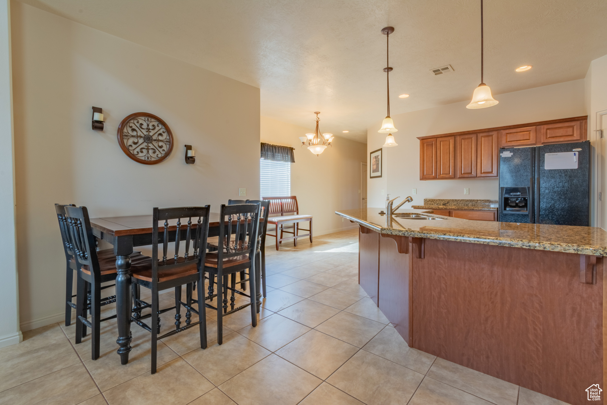 Kitchen featuring light stone counters, visible vents, hanging light fixtures, black fridge, and brown cabinetry