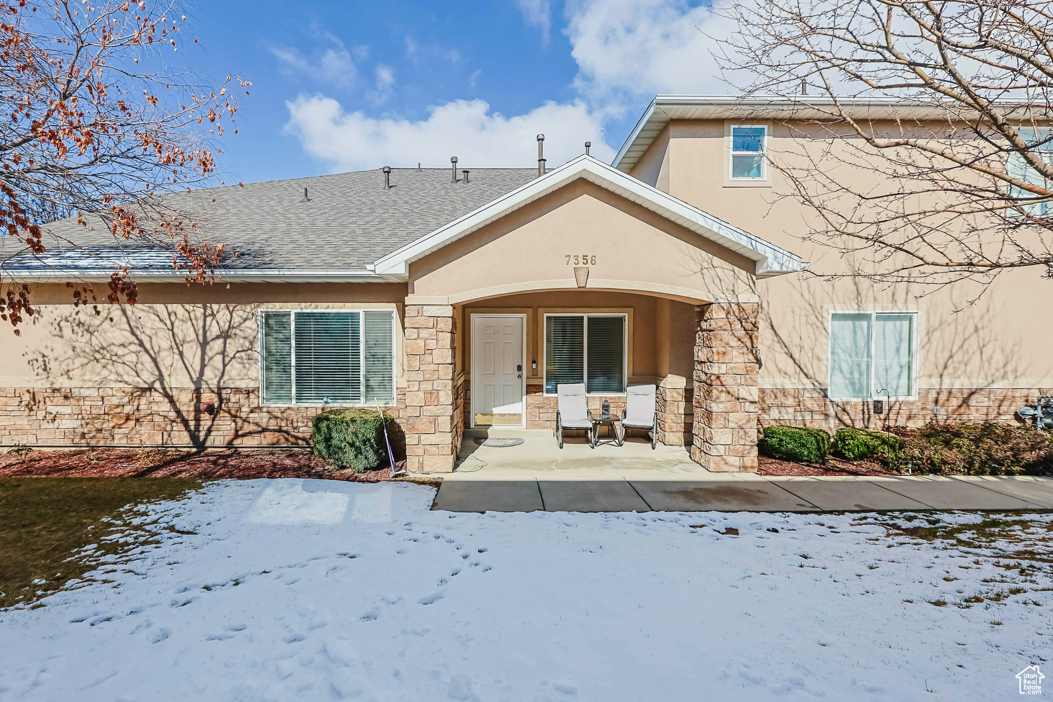 Snow covered back of property featuring stone siding, a patio, roof with shingles, and stucco siding