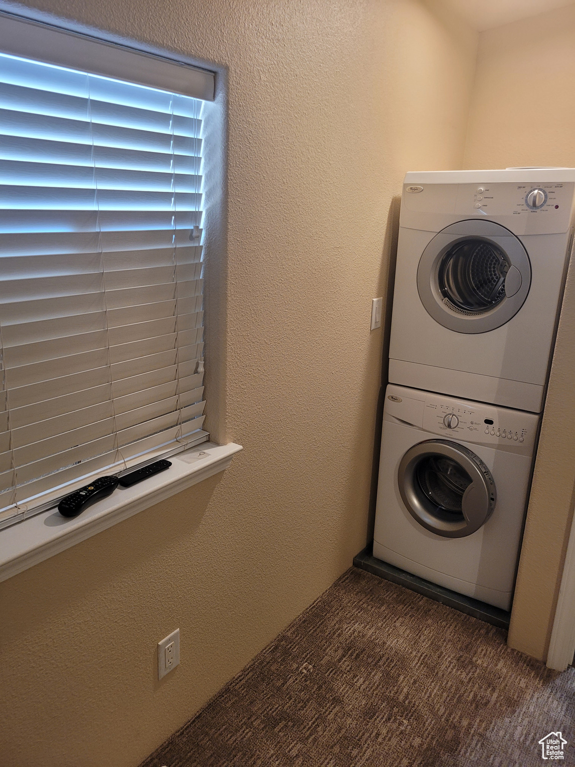 Clothes washing area featuring a textured wall, stacked washer and dryer, dark carpet, and laundry area