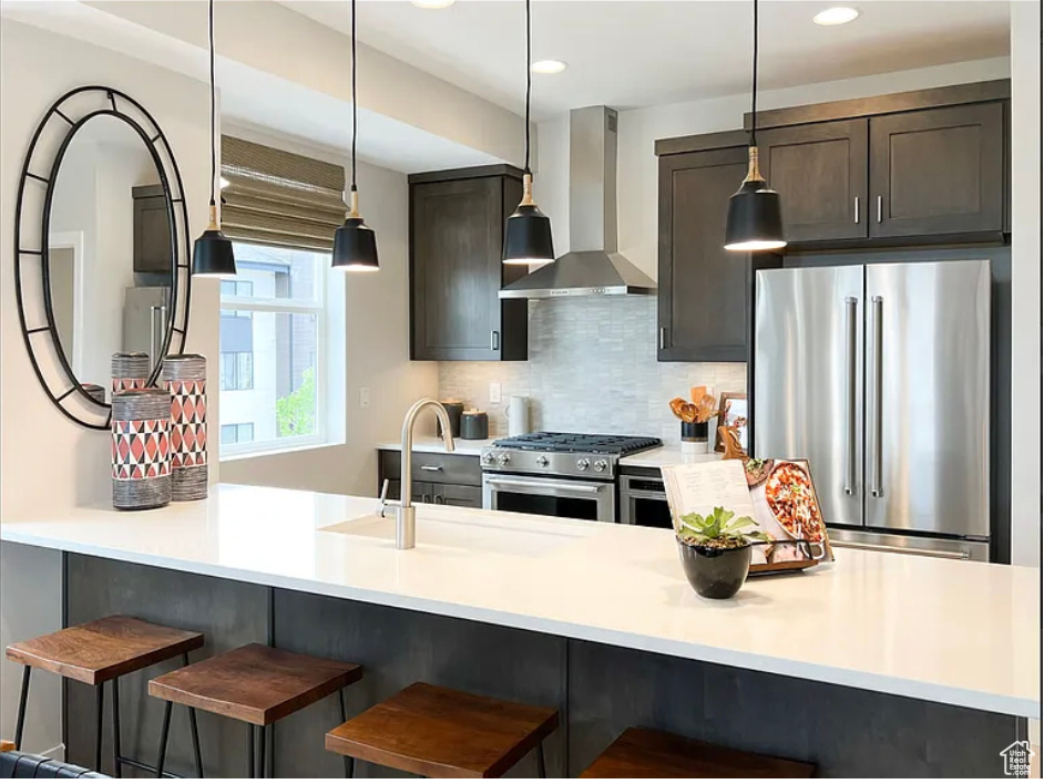 Kitchen featuring stainless steel appliances, dark brown cabinets, light countertops, wall chimney range hood, and a kitchen bar