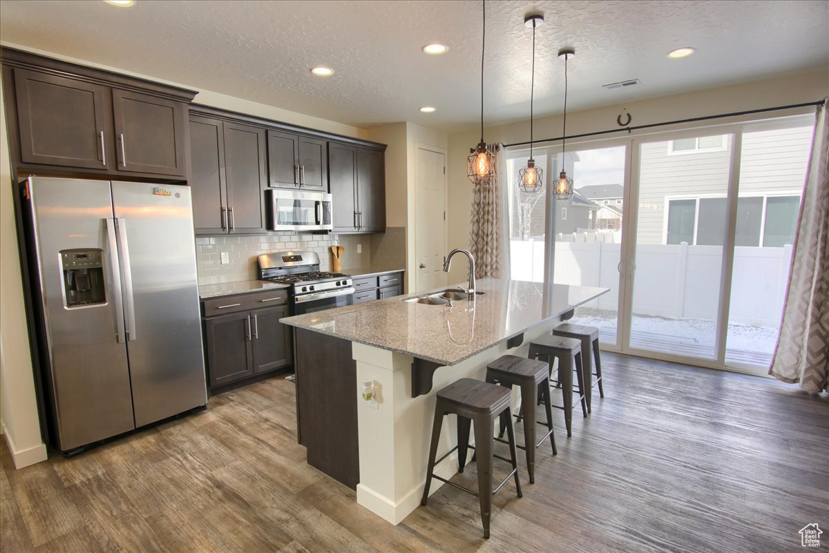Kitchen with a center island with sink, appliances with stainless steel finishes, light stone counters, a kitchen bar, and a sink