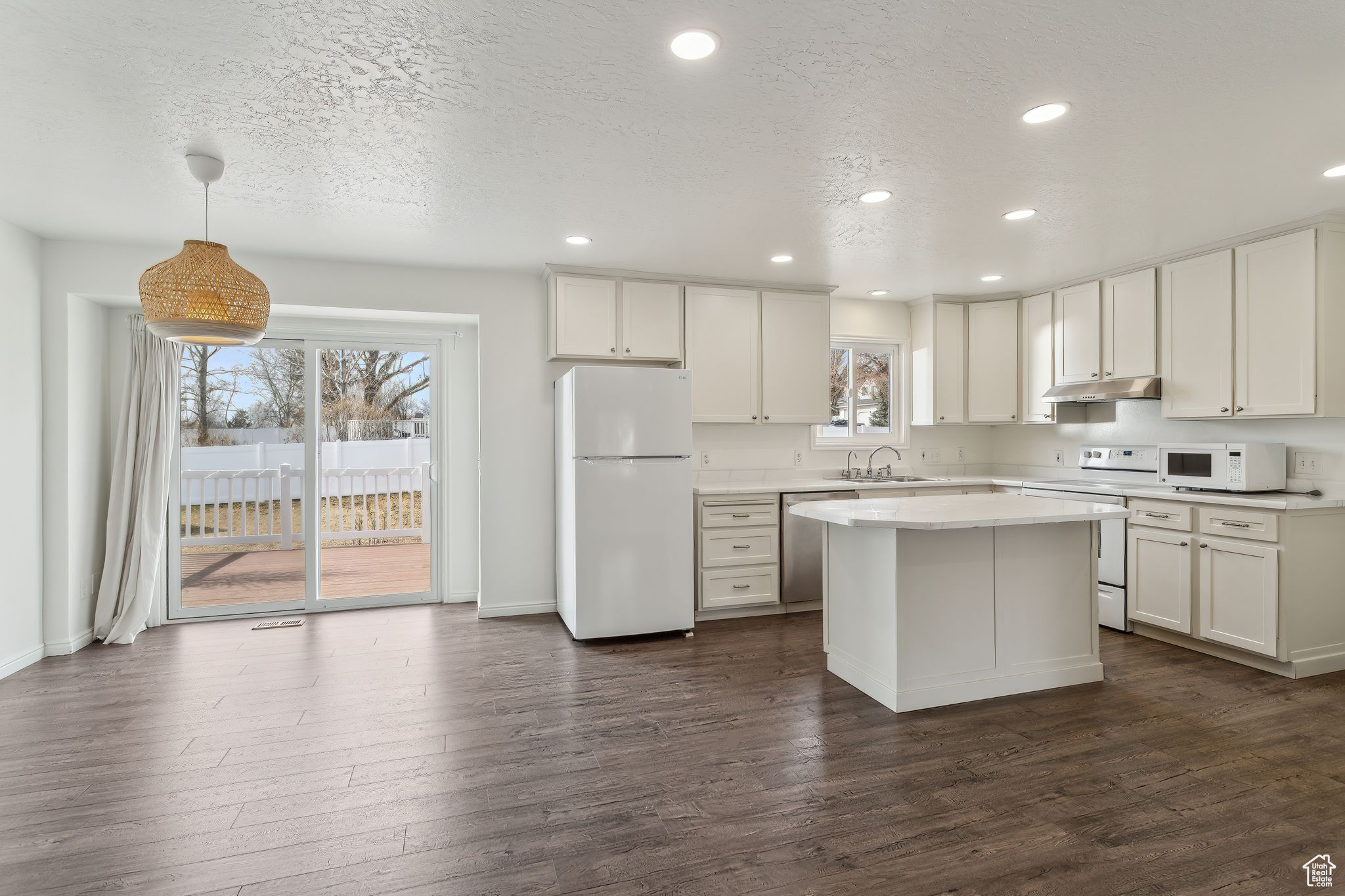 Kitchen featuring white appliances, decorative light fixtures, light countertops, under cabinet range hood, and white cabinetry