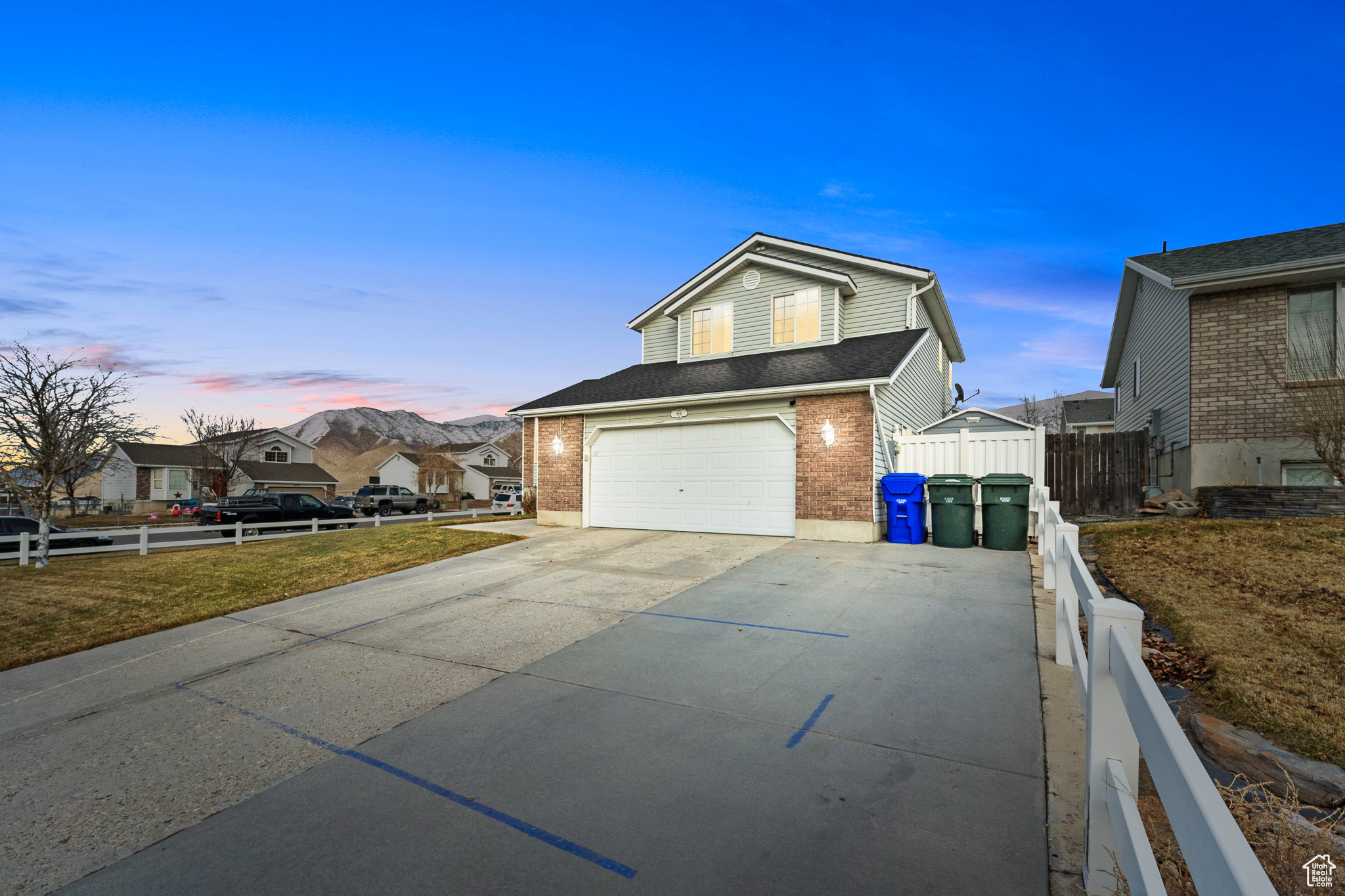 Property exterior at dusk with driveway, a residential view, an attached garage, fence, and brick siding