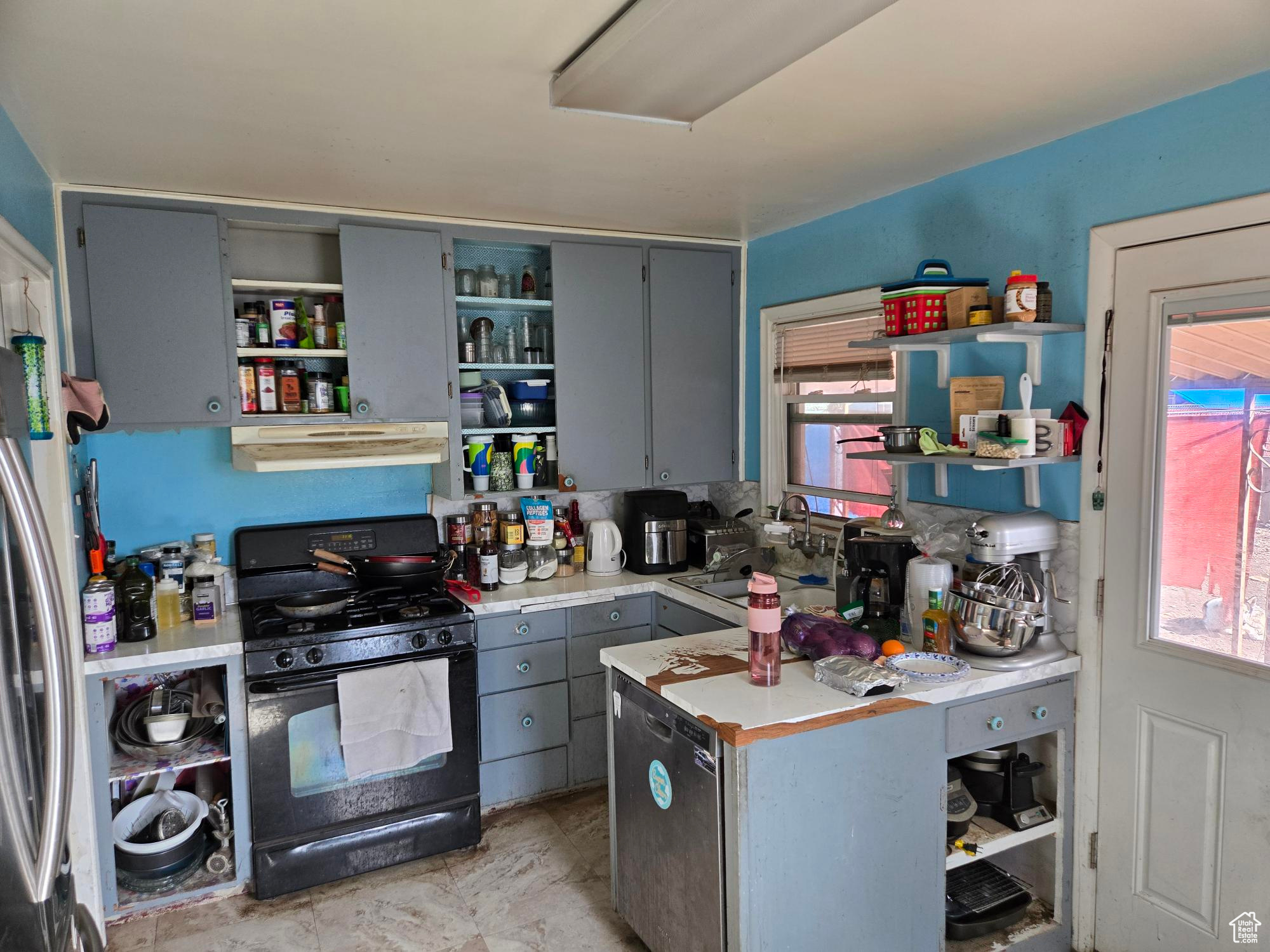 Kitchen featuring black gas range, under cabinet range hood, light countertops, freestanding refrigerator, and open shelves