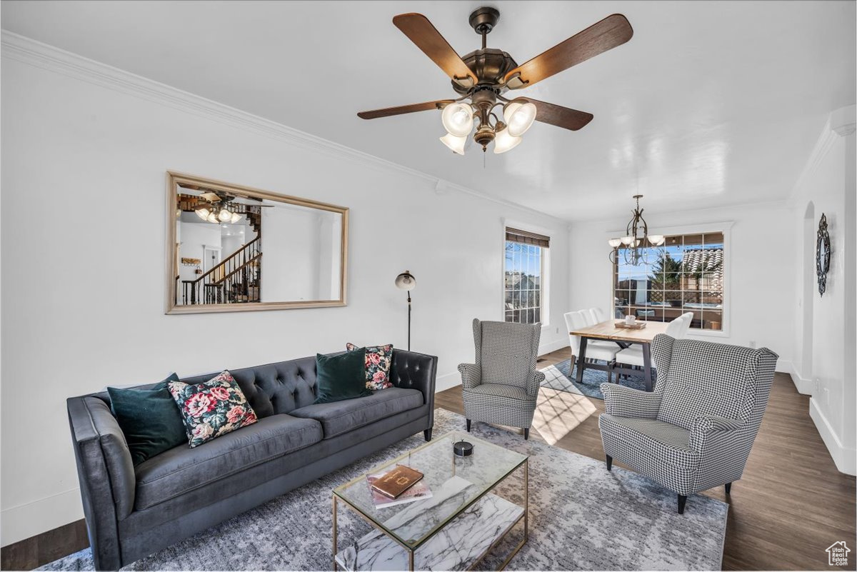 Living area with ceiling fan with notable chandelier, baseboards, ornamental molding, stairway, and dark wood-style floors