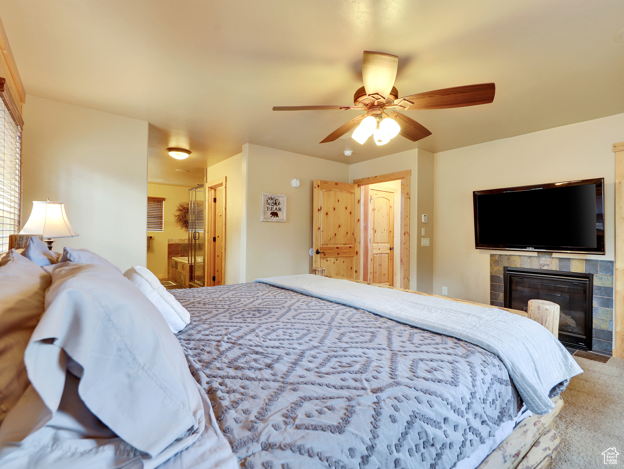 Carpeted bedroom featuring a tiled fireplace, ensuite bath, and ceiling fan