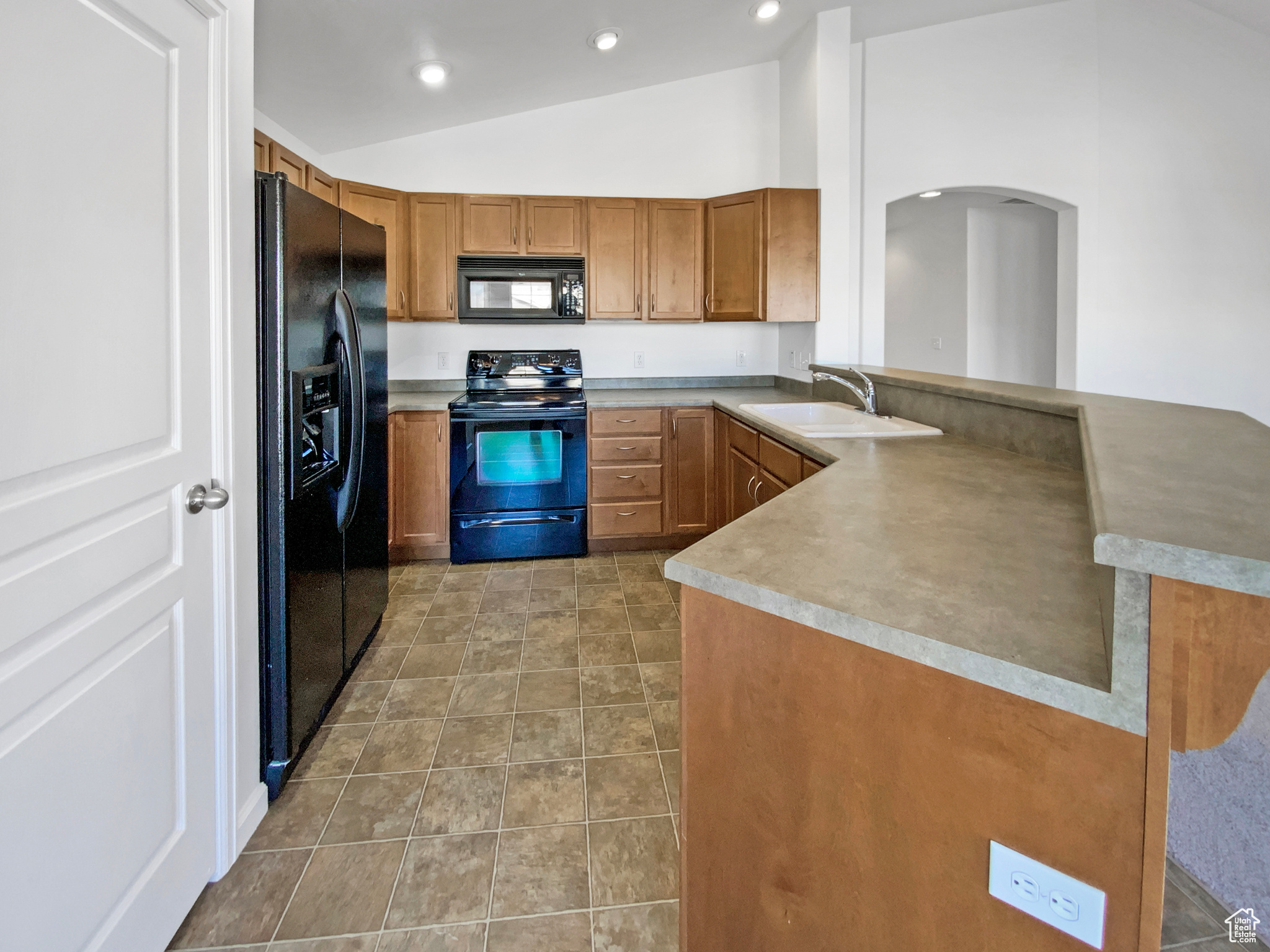 Kitchen with brown cabinetry, a peninsula, lofted ceiling, a sink, and black appliances