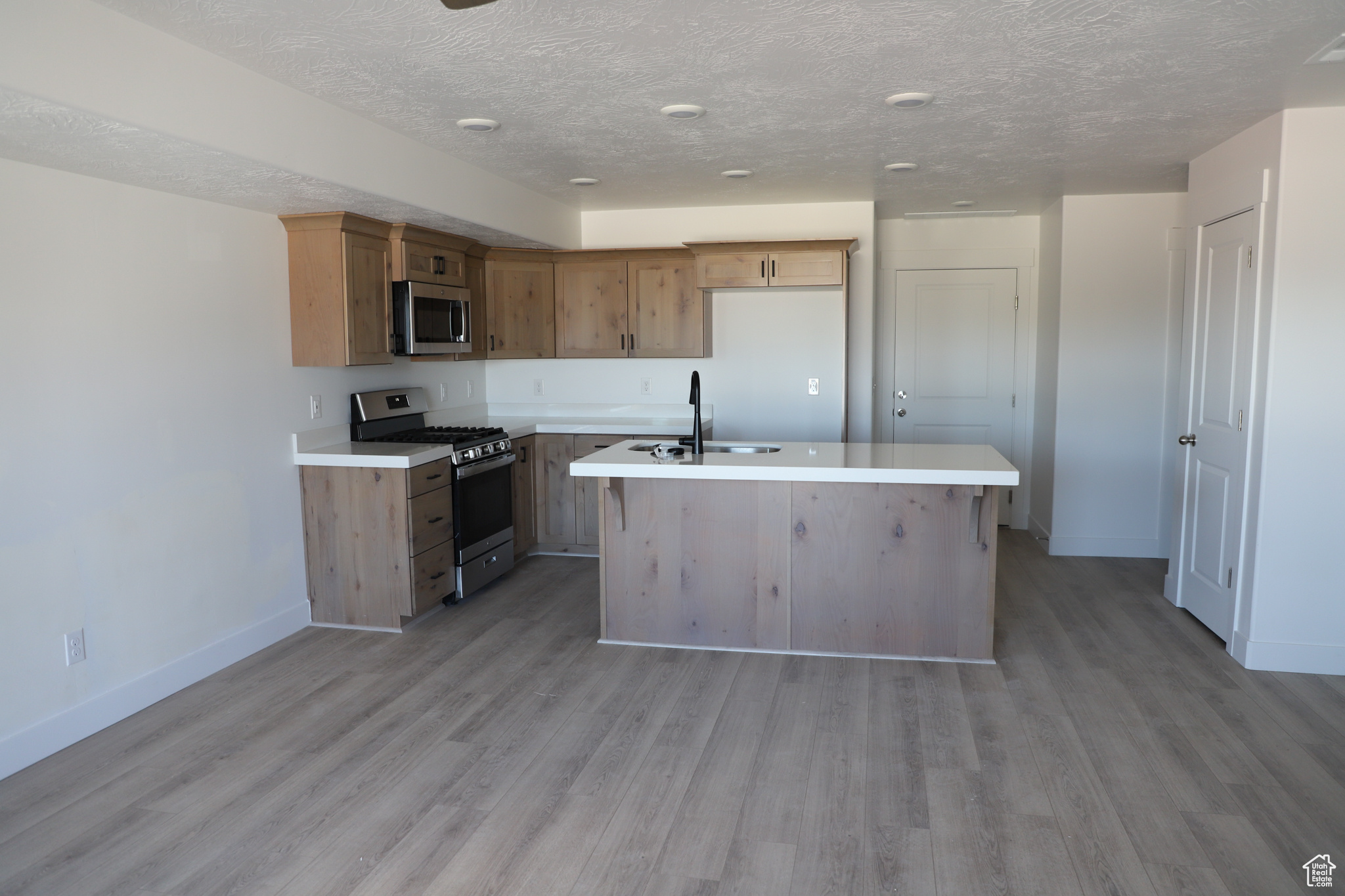 Kitchen with stainless steel appliances, an island with sink, light wood-style flooring, and light countertops