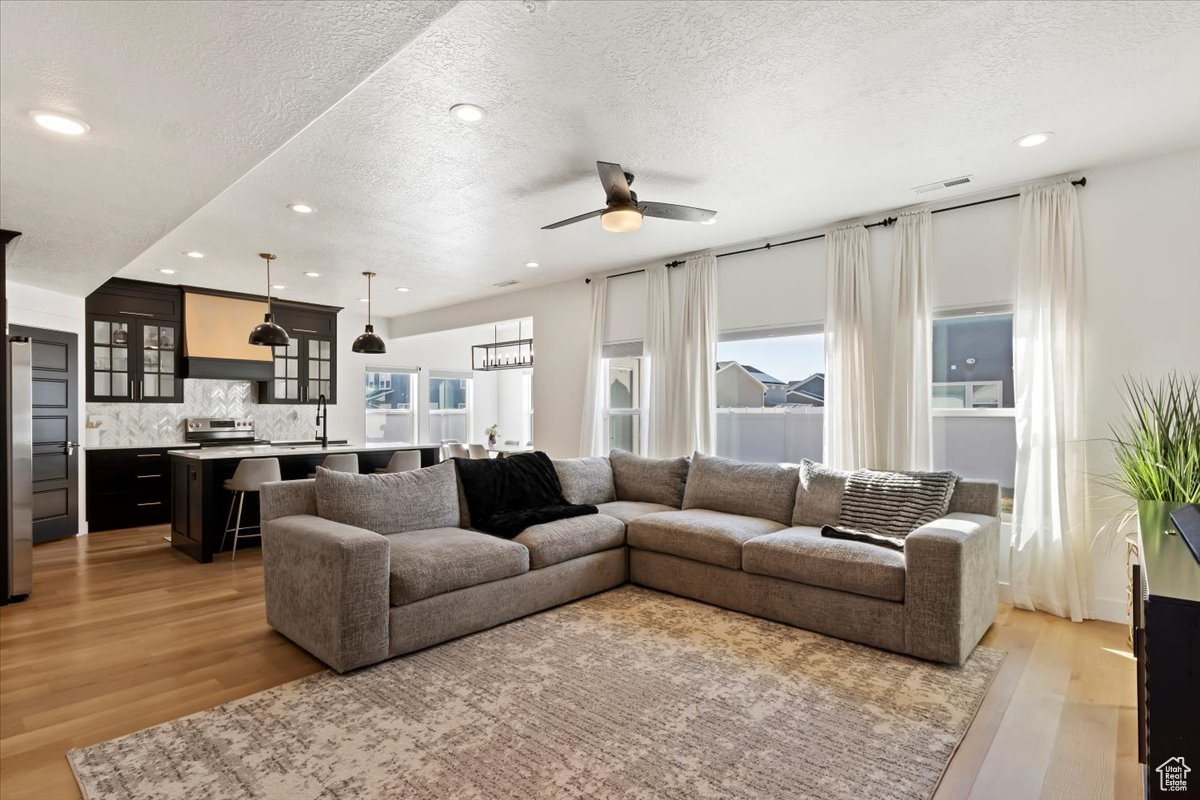 Living area with visible vents, light wood-type flooring, recessed lighting, ceiling fan with notable chandelier, and a textured ceiling