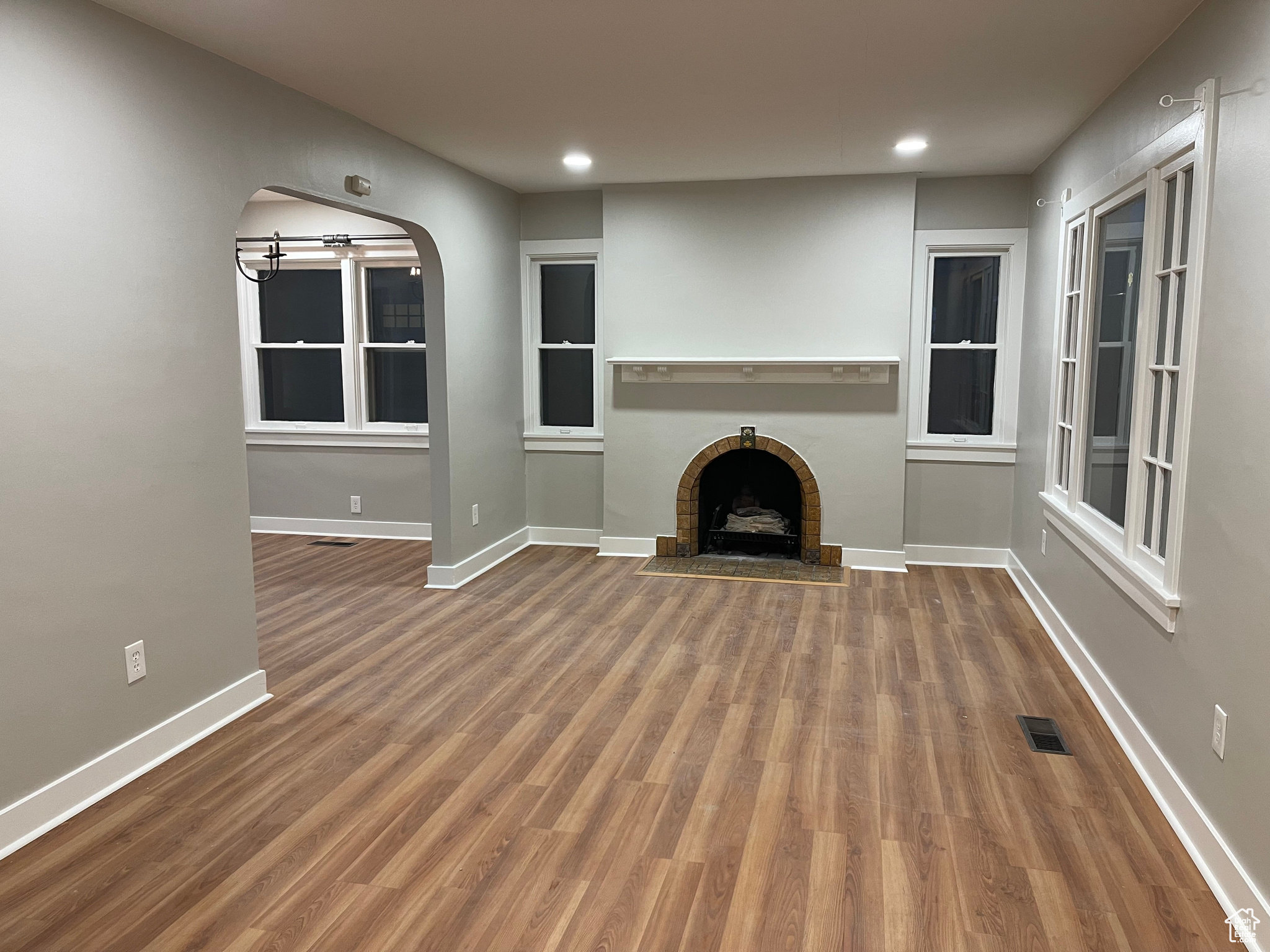 Unfurnished living room with arched walkways, visible vents, a brick fireplace, wood finished floors, and baseboards