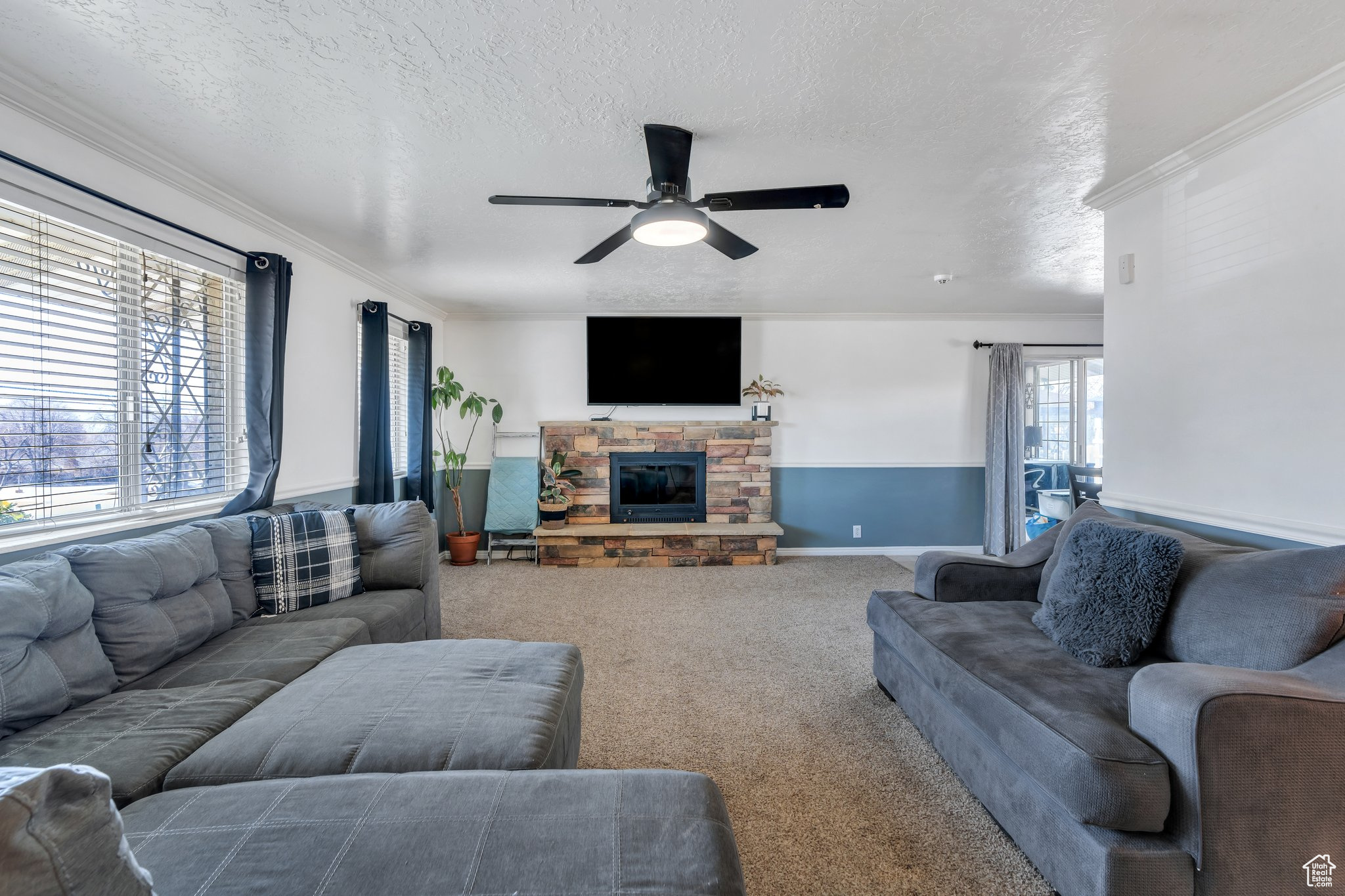 Carpeted living room with a stone fireplace, a textured ceiling, a ceiling fan, and ornamental molding