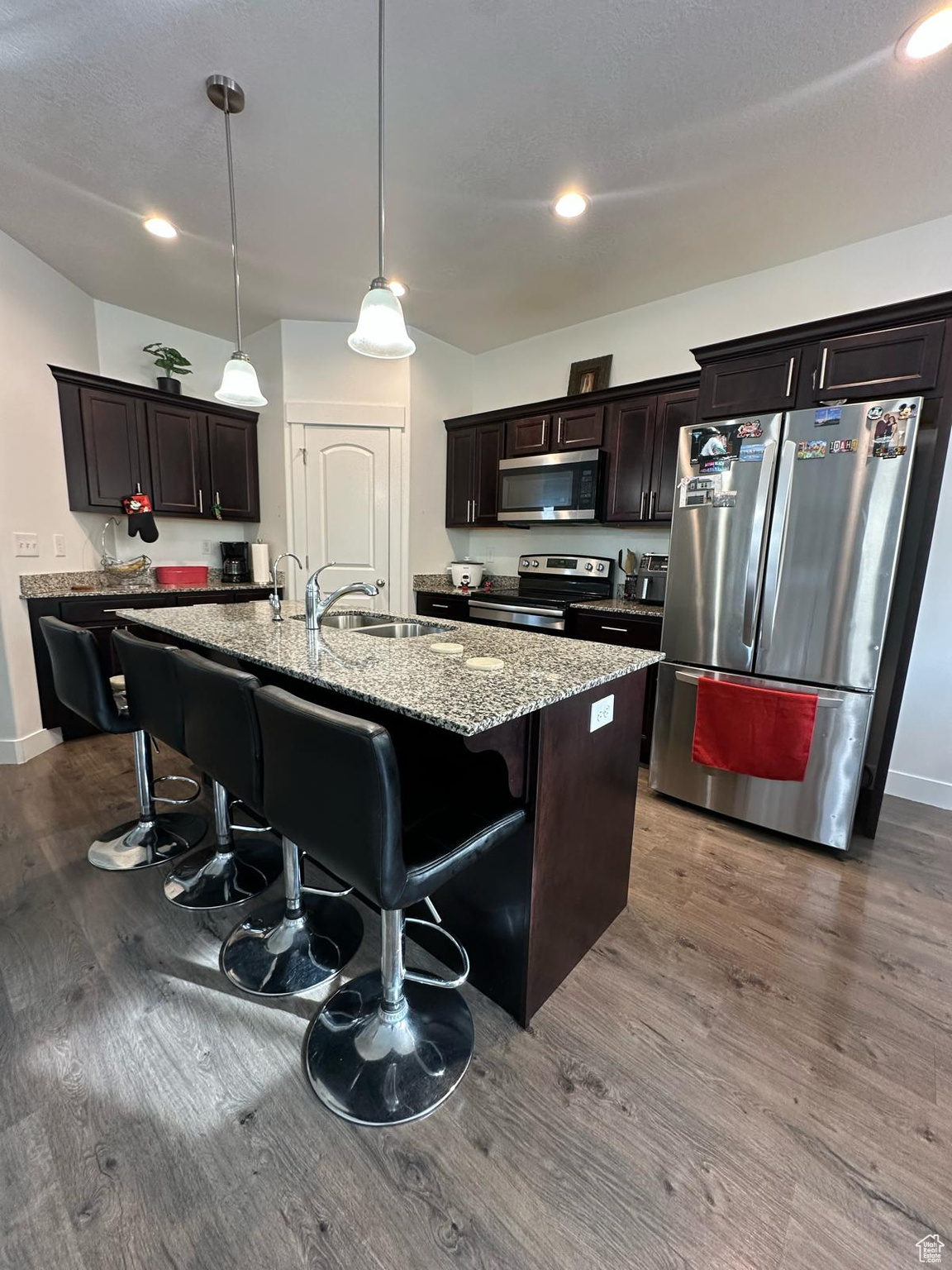 Kitchen featuring light stone counters, dark wood-type flooring, a kitchen island with sink, stainless steel appliances, and a sink