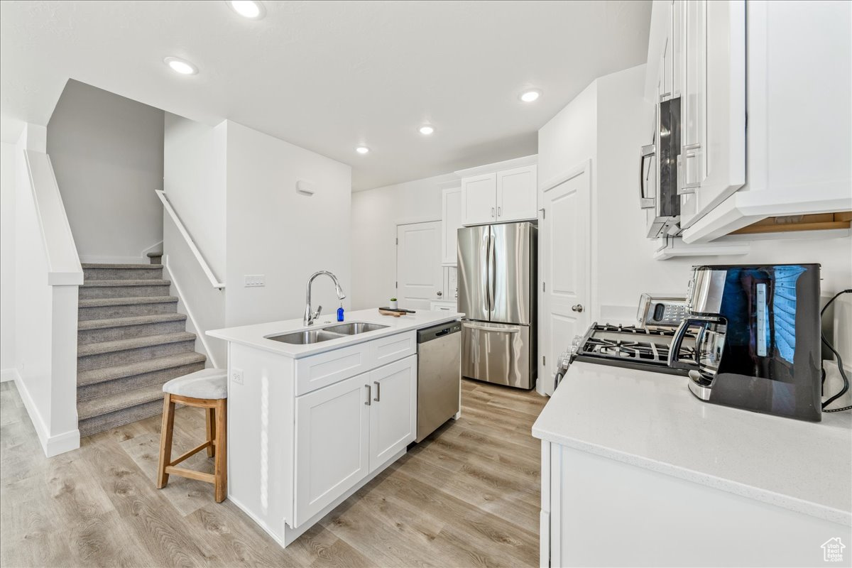Kitchen with light wood finished floors, a kitchen island with sink, a sink, white cabinets, and appliances with stainless steel finishes