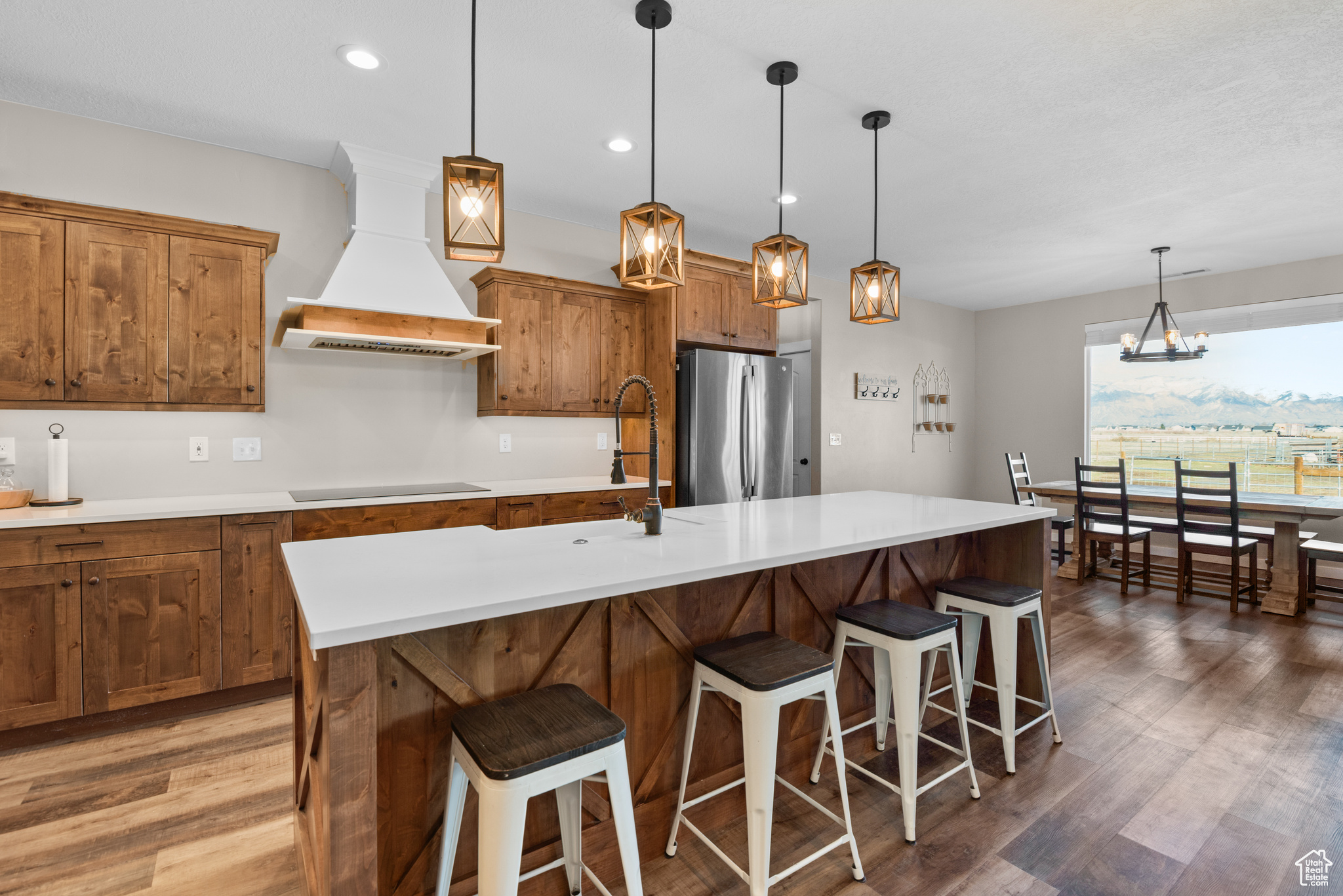 Kitchen with black electric stovetop, custom exhaust hood, brown cabinetry, freestanding refrigerator, and wood finished floors