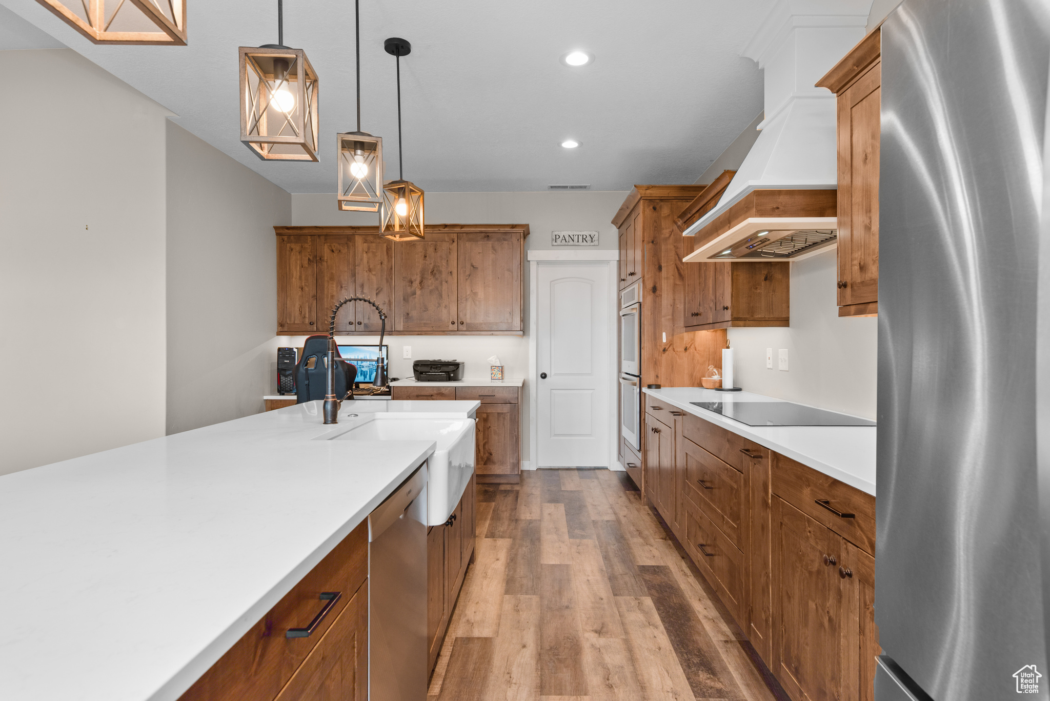 Kitchen featuring wood finished floors, appliances with stainless steel finishes, brown cabinetry, decorative light fixtures, and custom range hood