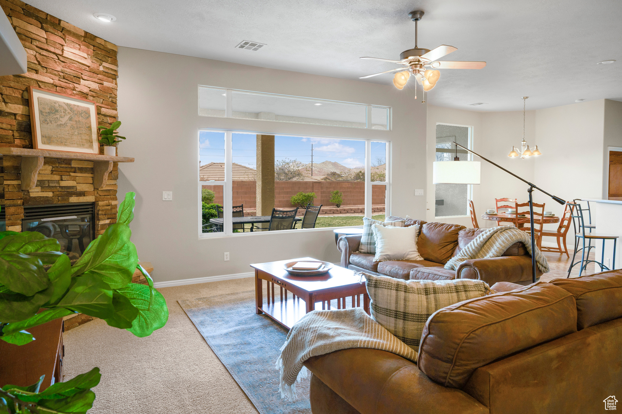 Carpeted living room featuring visible vents, ceiling fan with notable chandelier, a stone fireplace, and baseboards