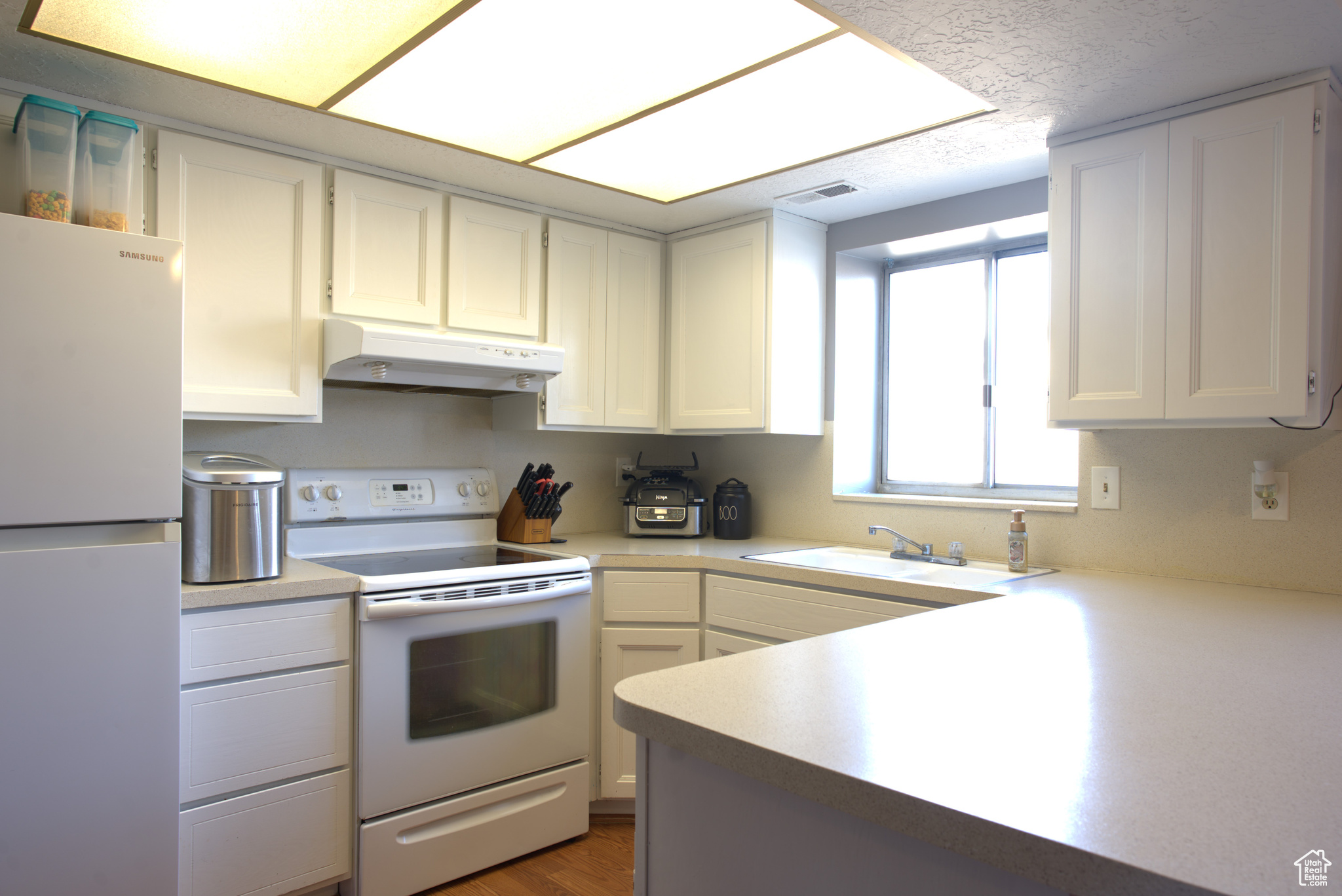 Kitchen featuring visible vents, under cabinet range hood, a sink, white appliances, and light countertops