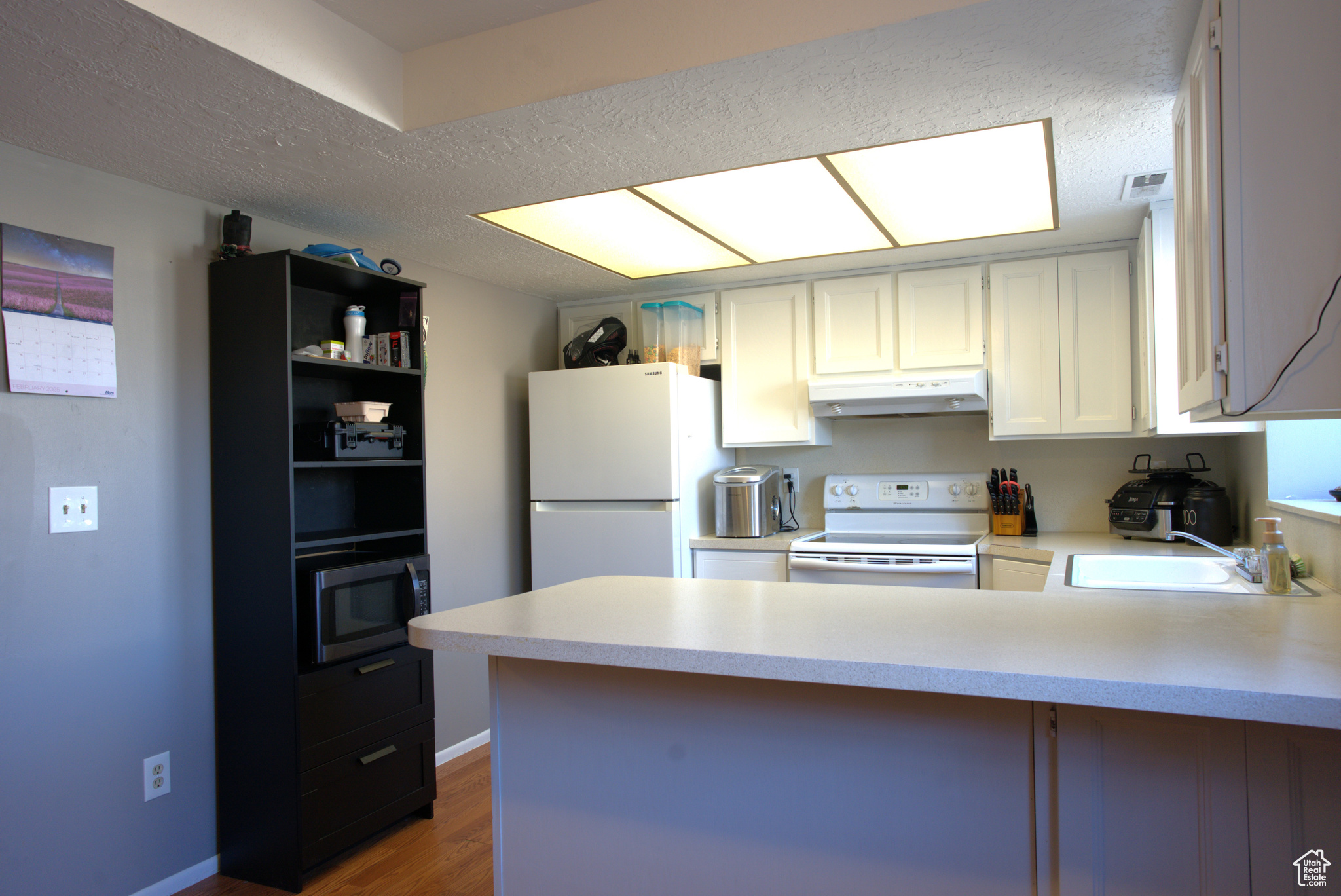 Kitchen featuring white appliances, a peninsula, a sink, light countertops, and under cabinet range hood