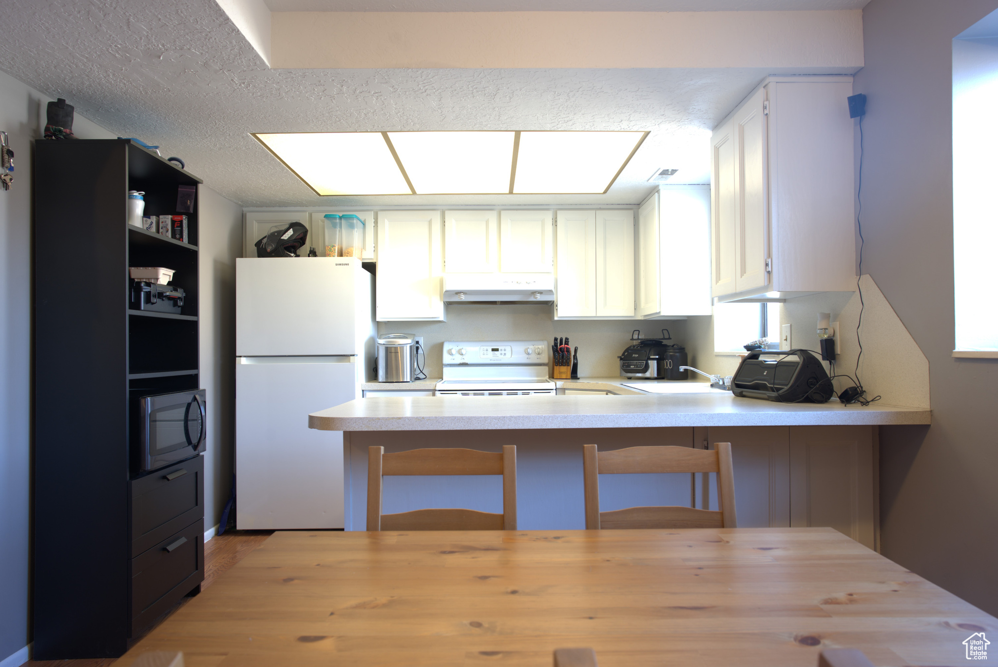 Kitchen with under cabinet range hood, white appliances, a peninsula, white cabinets, and light countertops