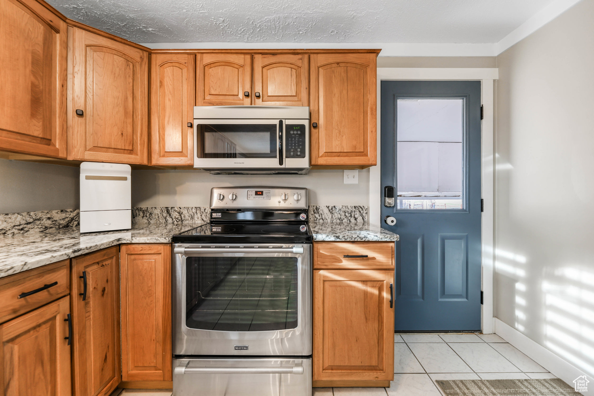 Kitchen with brown cabinets, light stone counters, appliances with stainless steel finishes, light tile patterned flooring, and baseboards