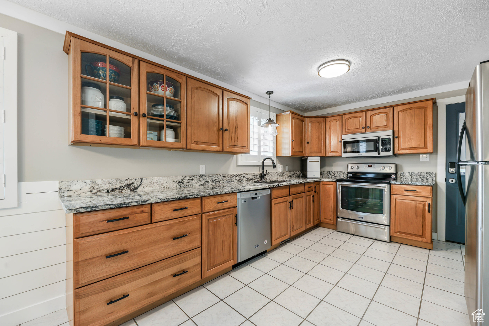 Kitchen with light stone countertops, brown cabinetry, a sink, glass insert cabinets, and appliances with stainless steel finishes