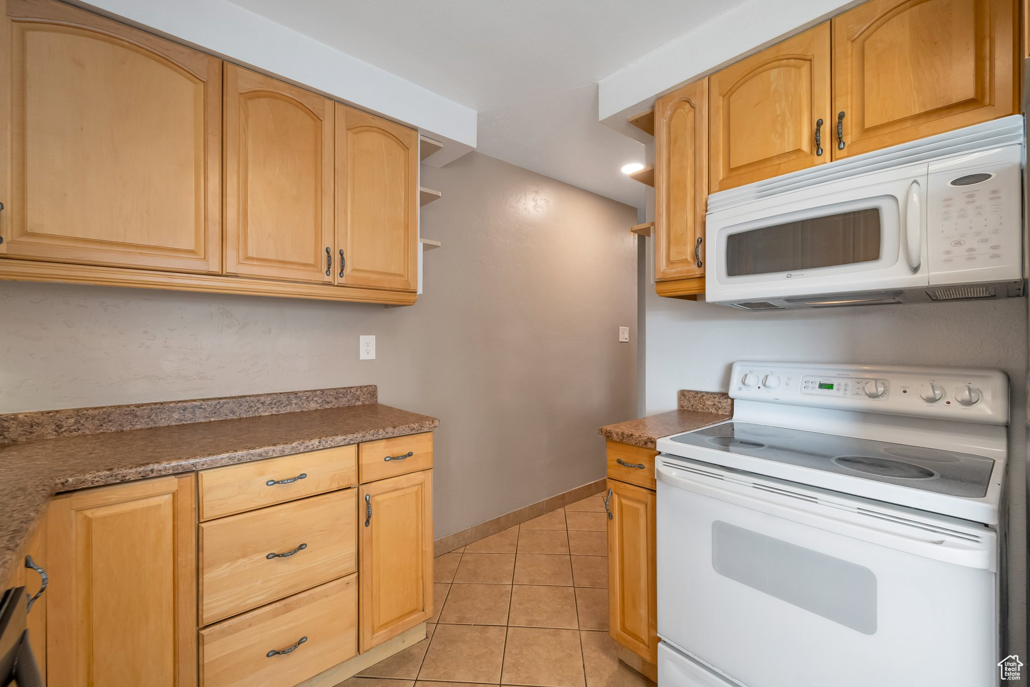 Kitchen featuring light tile patterned flooring, white appliances, dark countertops, and baseboards