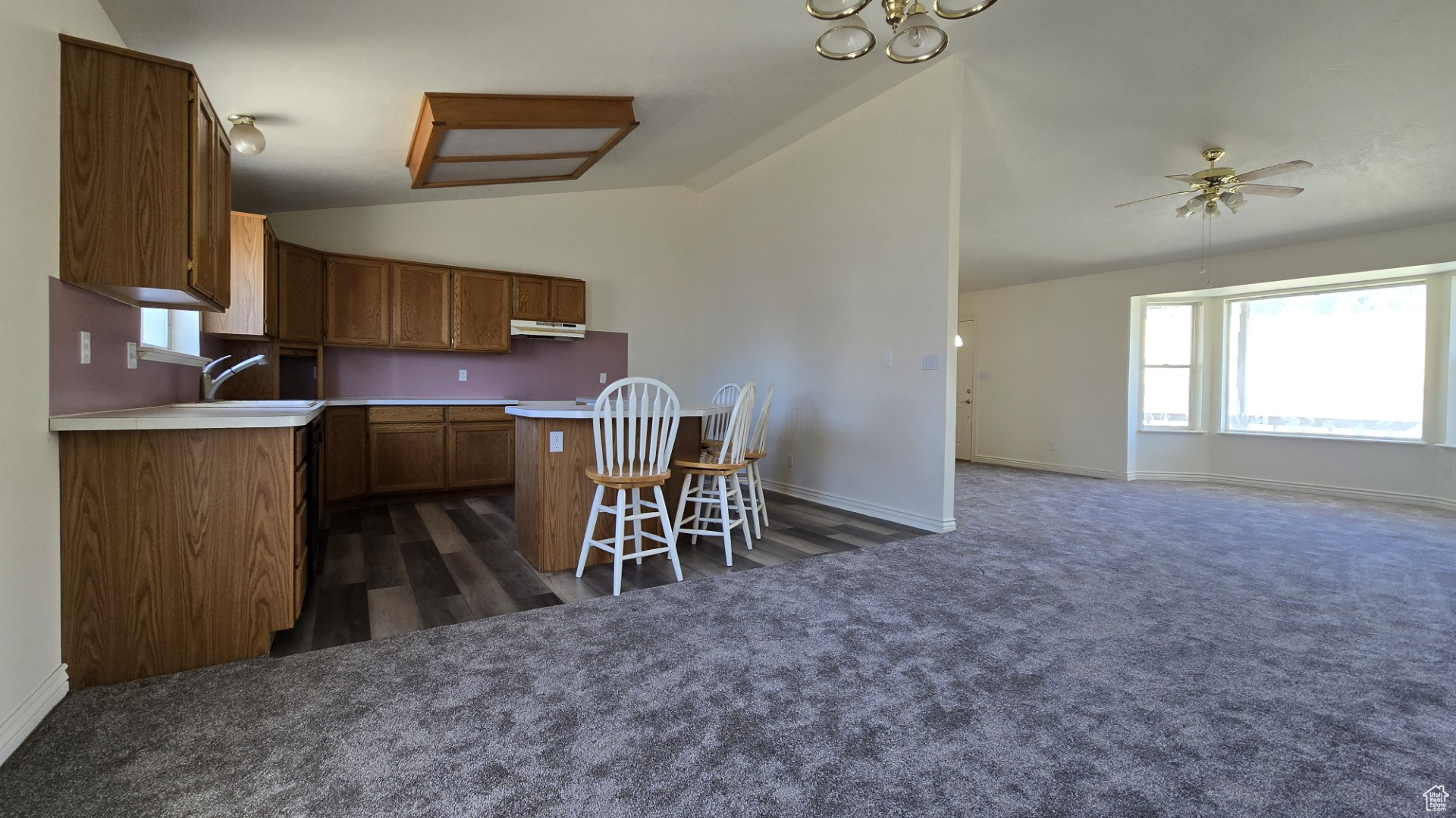 Kitchen featuring a breakfast bar area, brown cabinetry, lofted ceiling, under cabinet range hood, and dark colored carpet