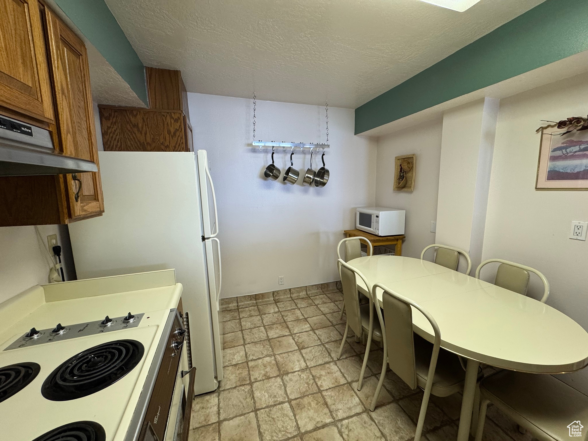 Kitchen with stone finish flooring, under cabinet range hood, brown cabinetry, white appliances, and a textured ceiling