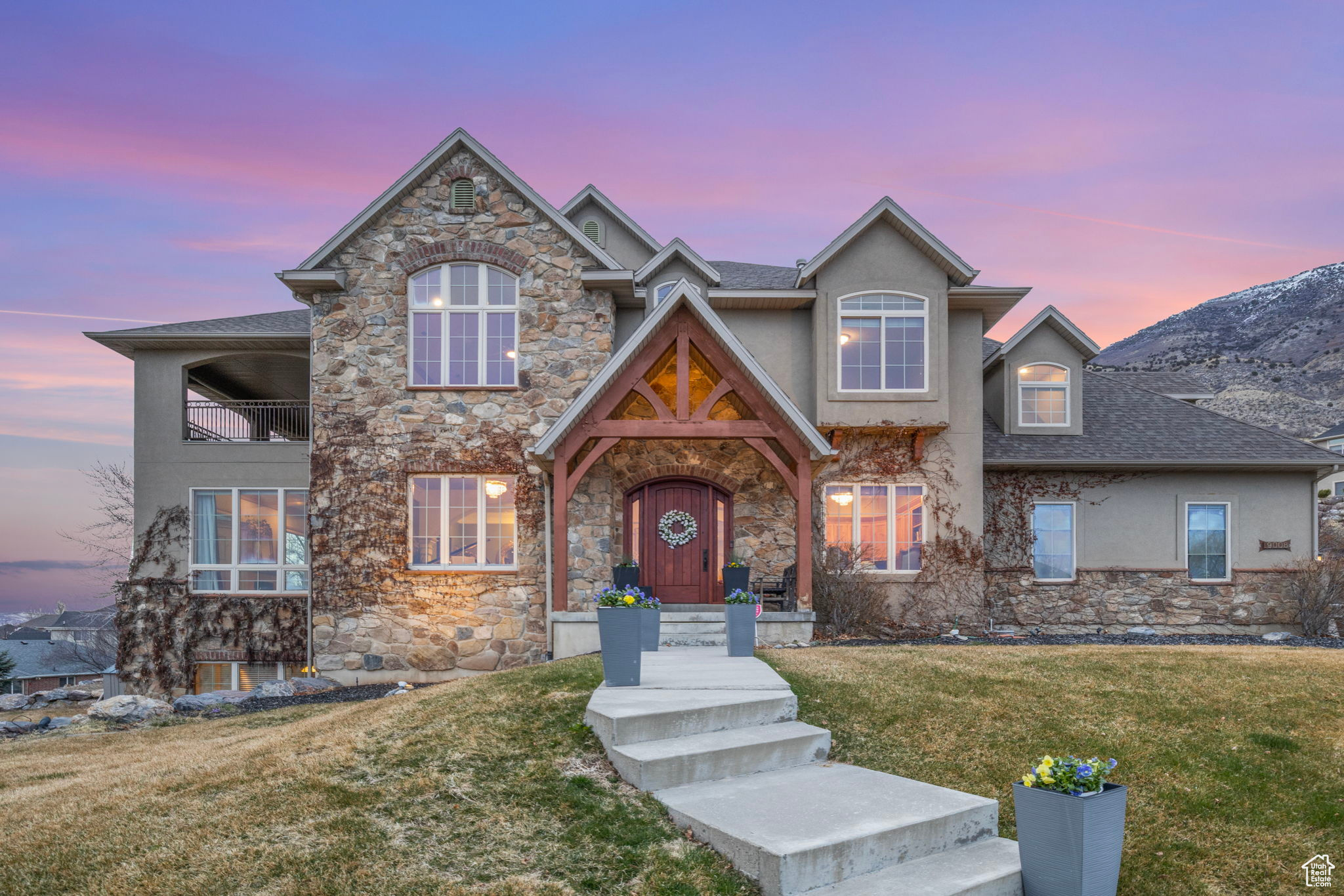 Stone-and-siding home with a timber-framed entry, front steps, and a snow-dusted mountain backdrop at dusk.