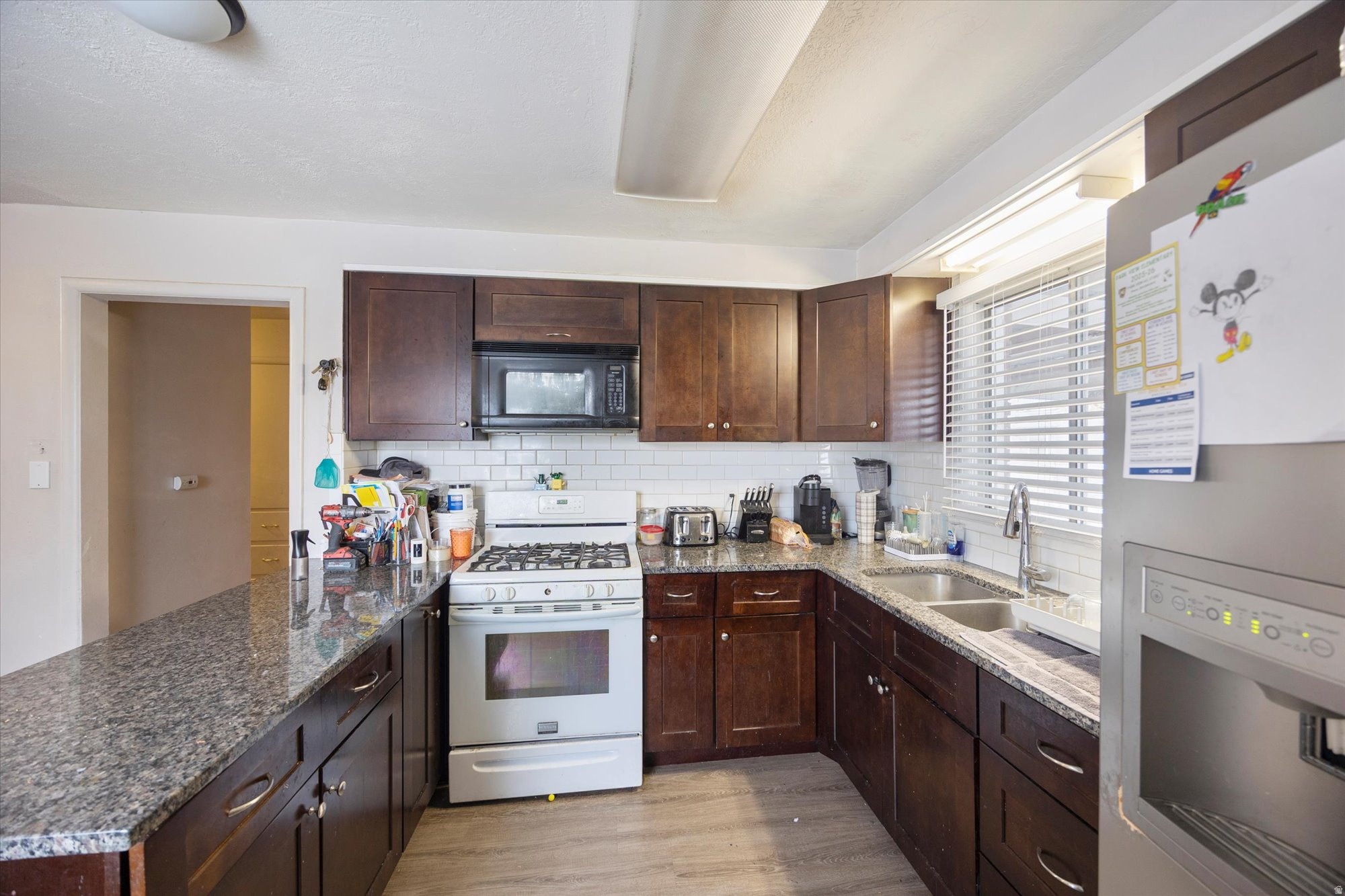 Kitchen featuring white gas stove, fridge with ice dispenser, black microwave, dark brown cabinets, and decorative backsplash