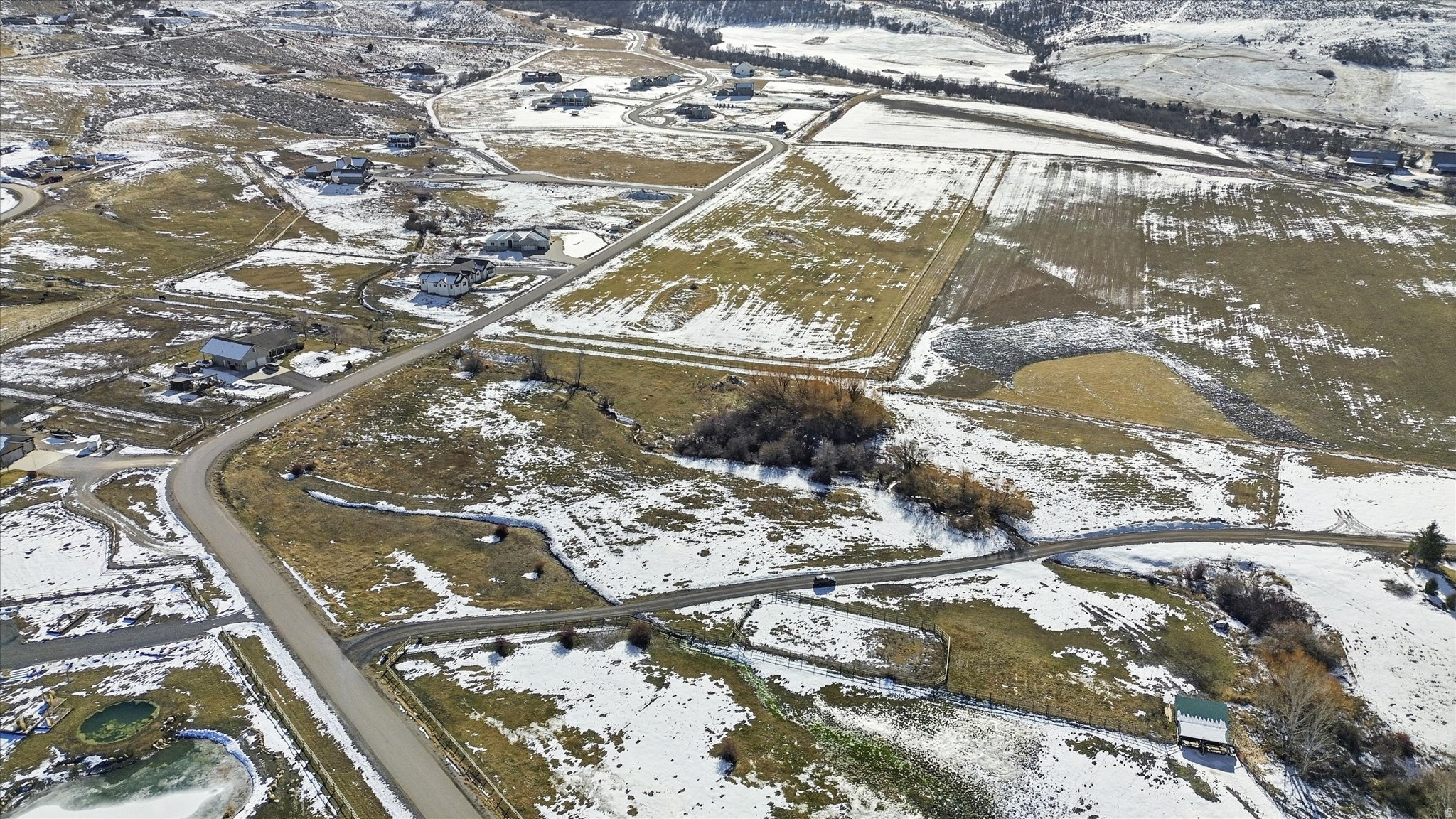 Expansive aerial view of a property featuring snow-dusted terrain, scattered residential structures, varied ground cover, winding roadways, and distant hills