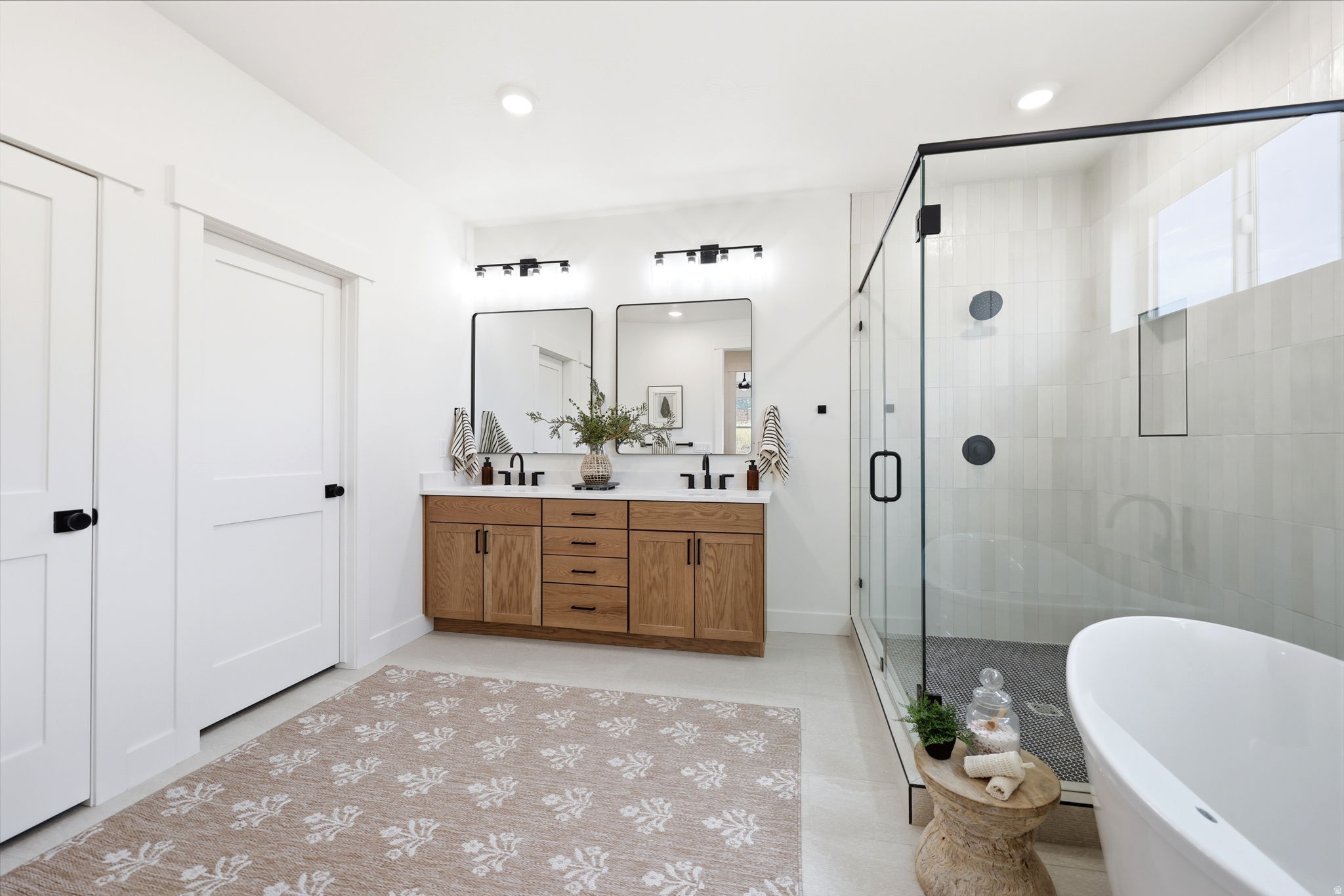 Modern bathroom featuring a dual vanity with wood-finish cabinetry, matte black fixtures, a spacious glass-enclosed shower, a freestanding soaking tub, and plank-style flooring