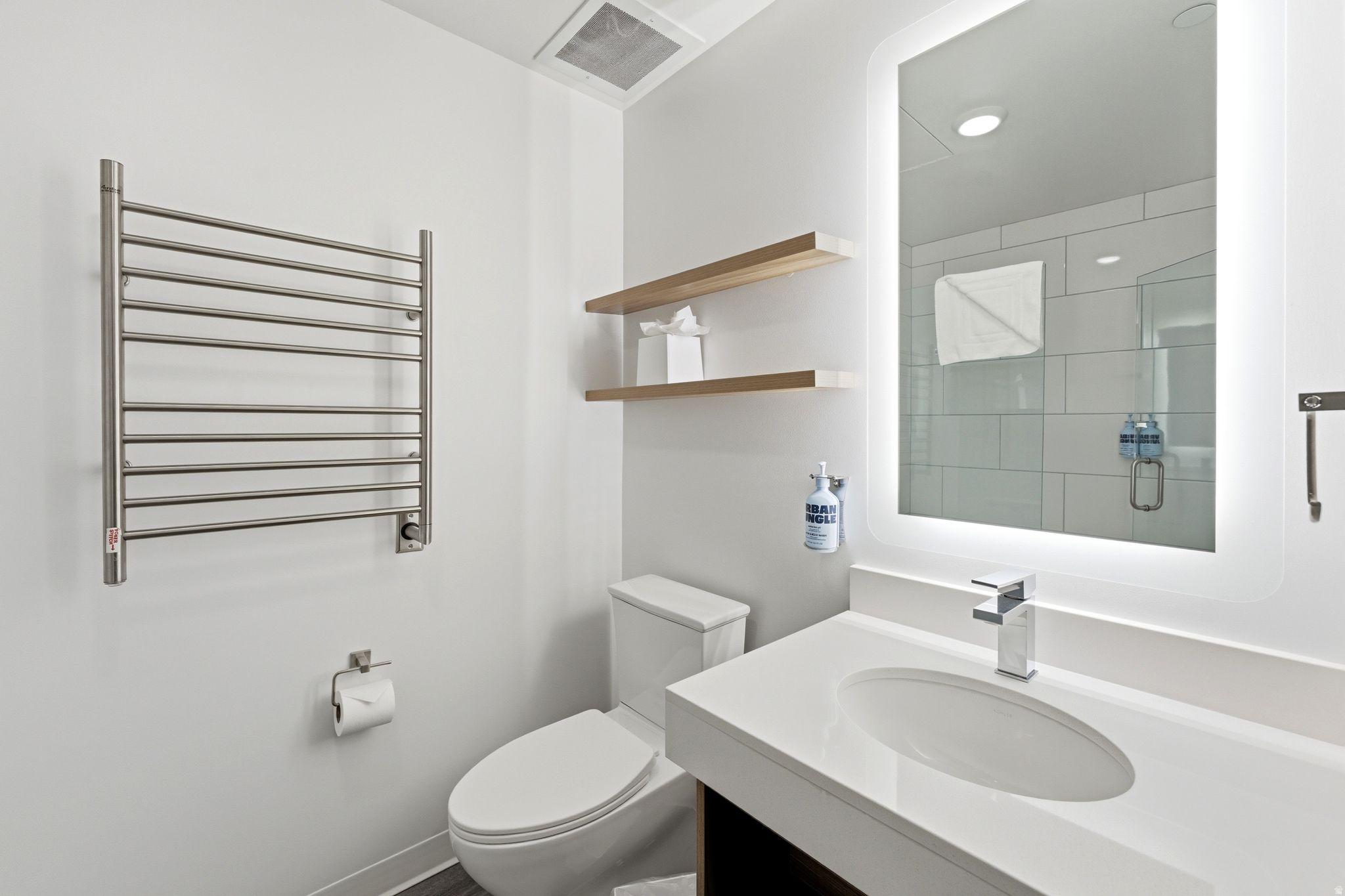 Modern bathroom featuring a vanity with an integrated sink, illuminated mirror, wall-mounted towel warmer, and floating wood-finish shelving