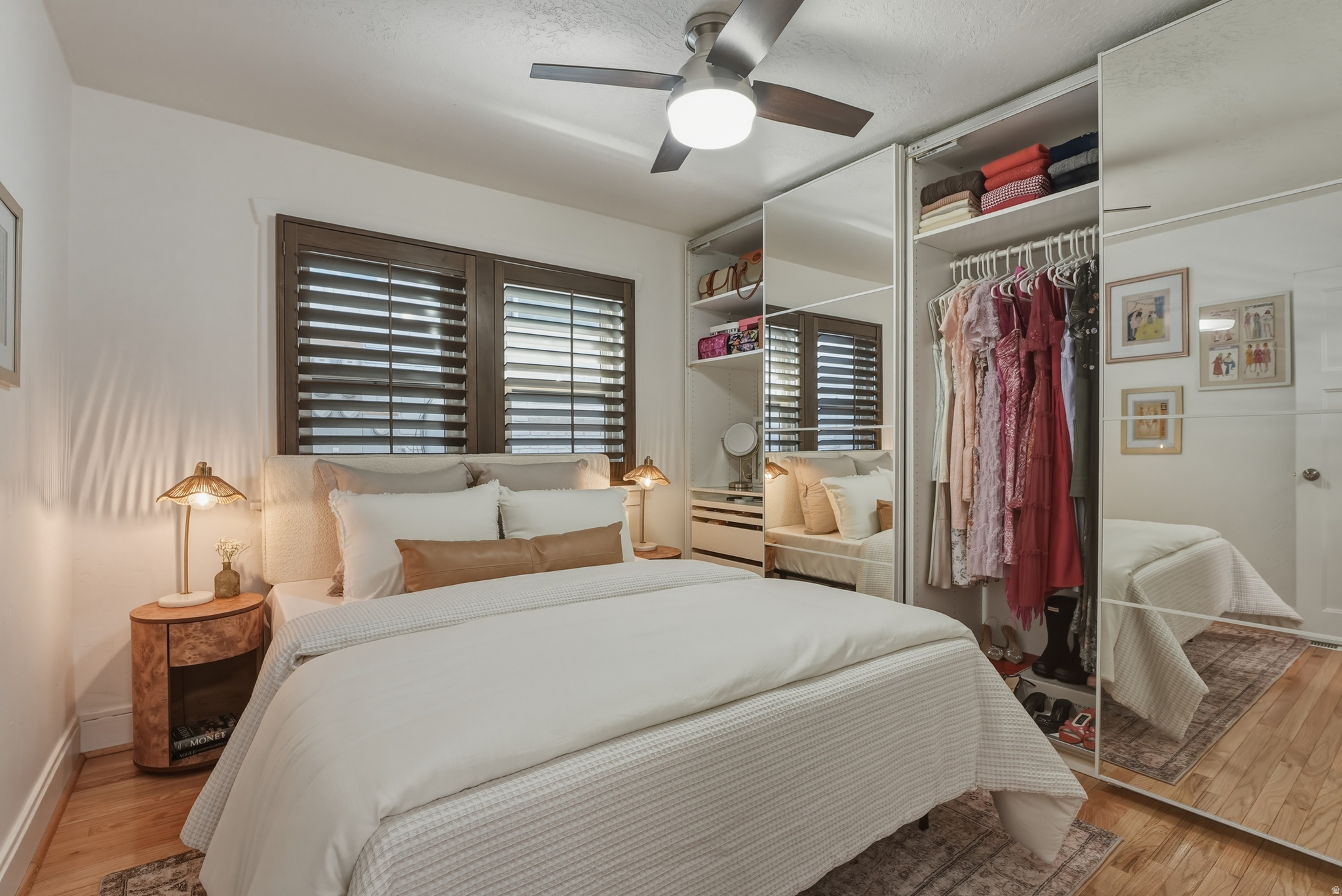 Bedroom featuring wood-finish flooring, dark wood window frames with plantation shutters, a ceiling fan, and mirrored wardrobe doors