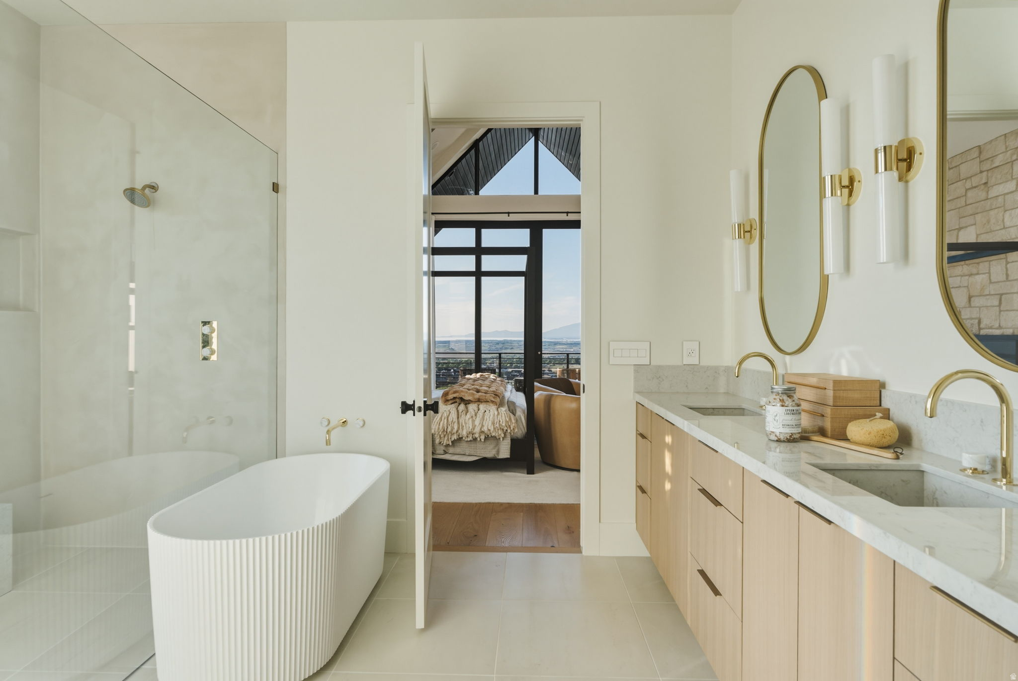 Contemporary bathroom featuring a fluted freestanding tub, a glass-enclosed shower, a double vanity with light wood-finish cabinetry, a light stone-finish countertop, and gold-finish fixtures