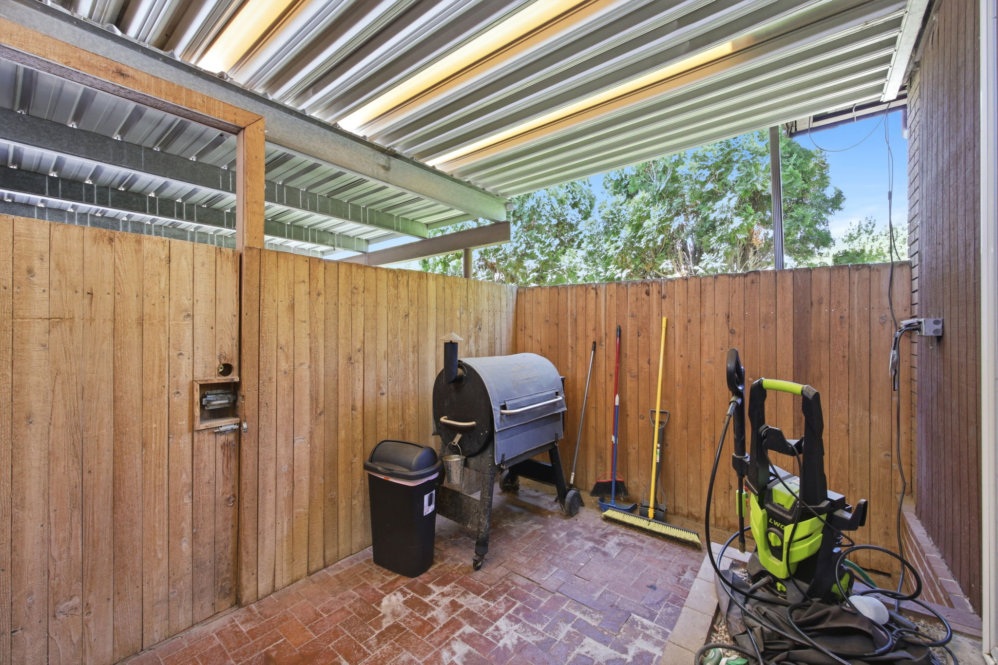 Covered outdoor patio area featuring a corrugated metal roof, brick paver flooring, and natural wood privacy fencing