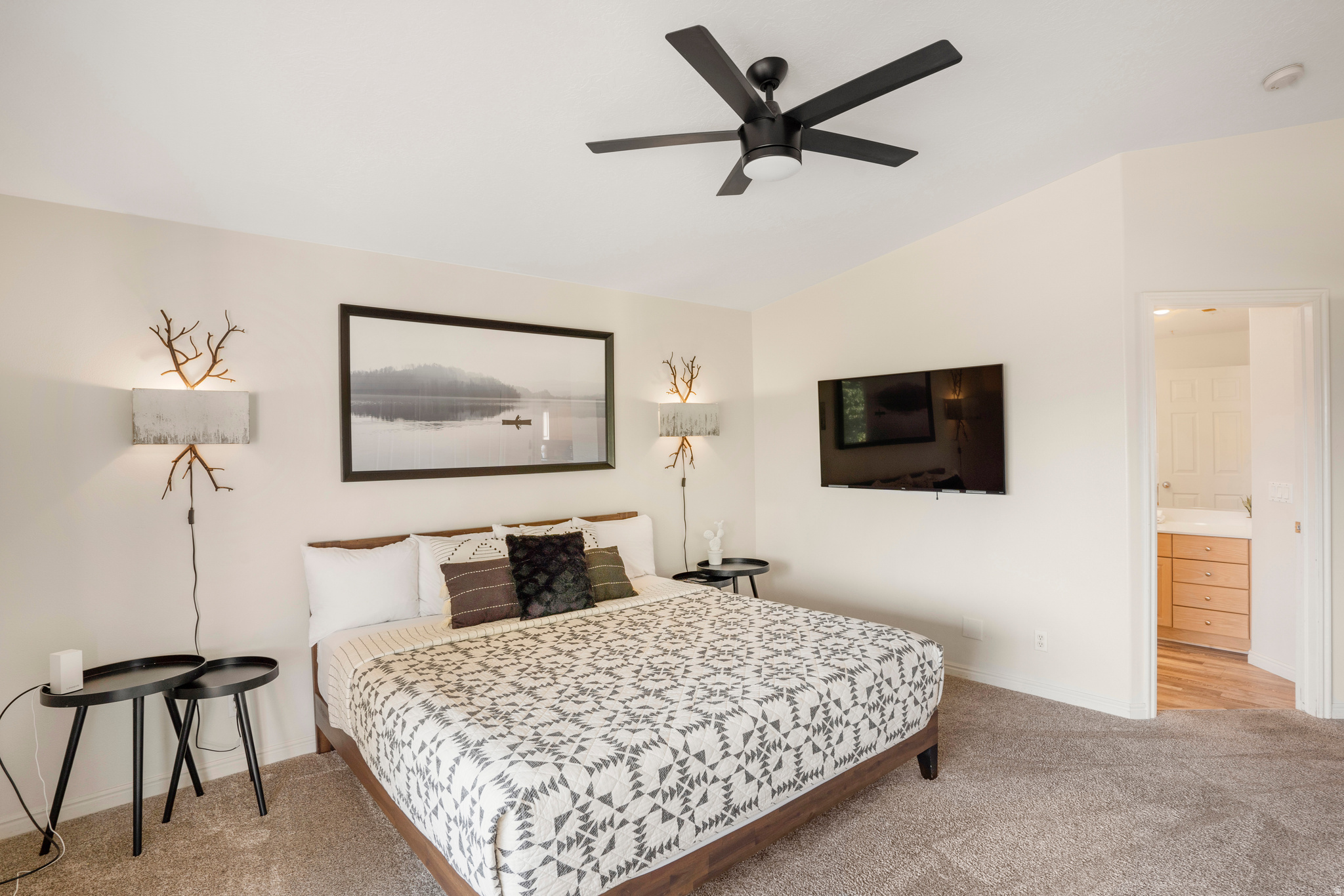 Carpeted bedroom featuring a ceiling fan with integrated lighting, wall-mounted television, and a doorway leading to an adjoining bathroom with wood-finish flooring and a vanity