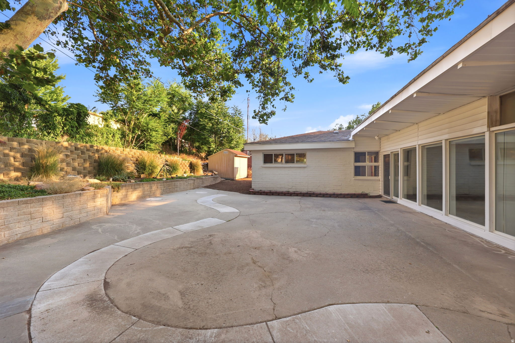 Extensive paved patio with a decorative curved inlay, a multi-tiered retaining wall with landscaping, a detached storage shed, and large floor-to-ceiling windows along the exterior wall