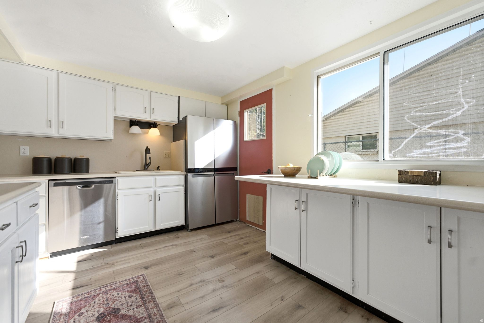 Bright, updated kitchen featuring white cabinetry, stainless steel appliances including a dishwasher and refrigerator, wood-finish flooring, and abundant natural light from large windows